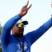 Palo Duro - Eric Mims 1 | Press Pass Sports Athlete in a blue long-sleeve shirt raises a two-finger victory sign with one hand, wearing a cap, sunglasses, and a lanyard outdoors.