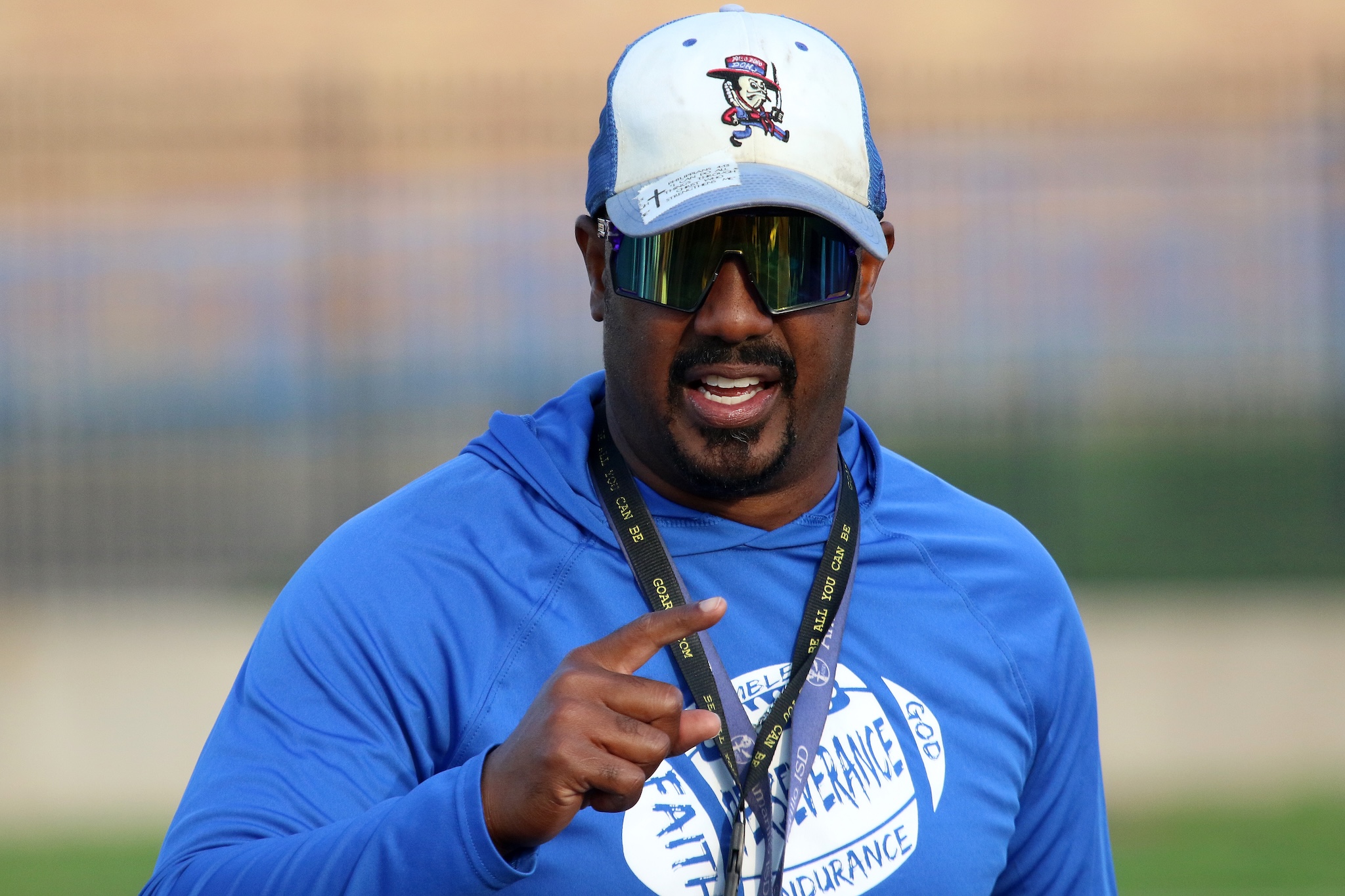 African American man outdoors in a blue shirt with a white cap and mirrored sunglasses, pointing toward the camera with a lanyard around his neck.