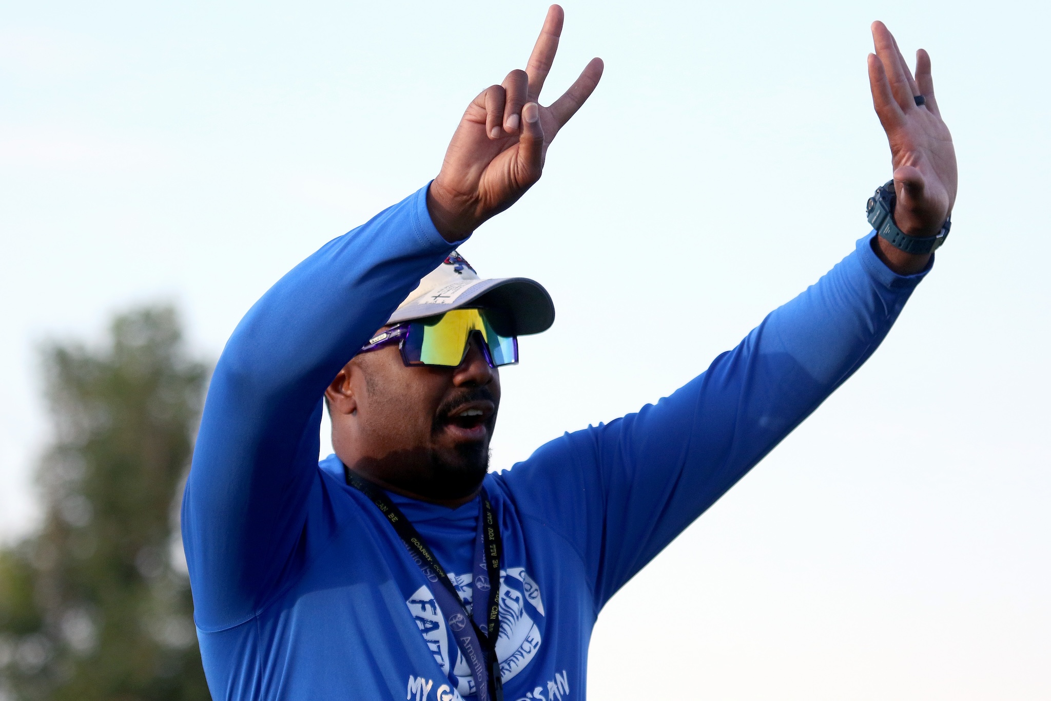 Athlete in a blue shirt and visor raises one hand in a peace sign while the other is raised in celebration outdoors.