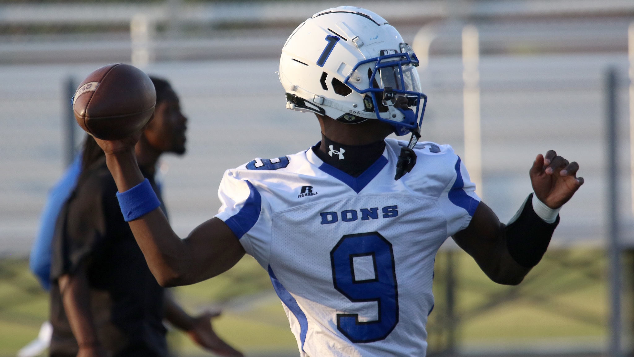 Quaterback in white and blue jersey (DONS) prepares to throw a football during a game, with blue helmet and focus forward.