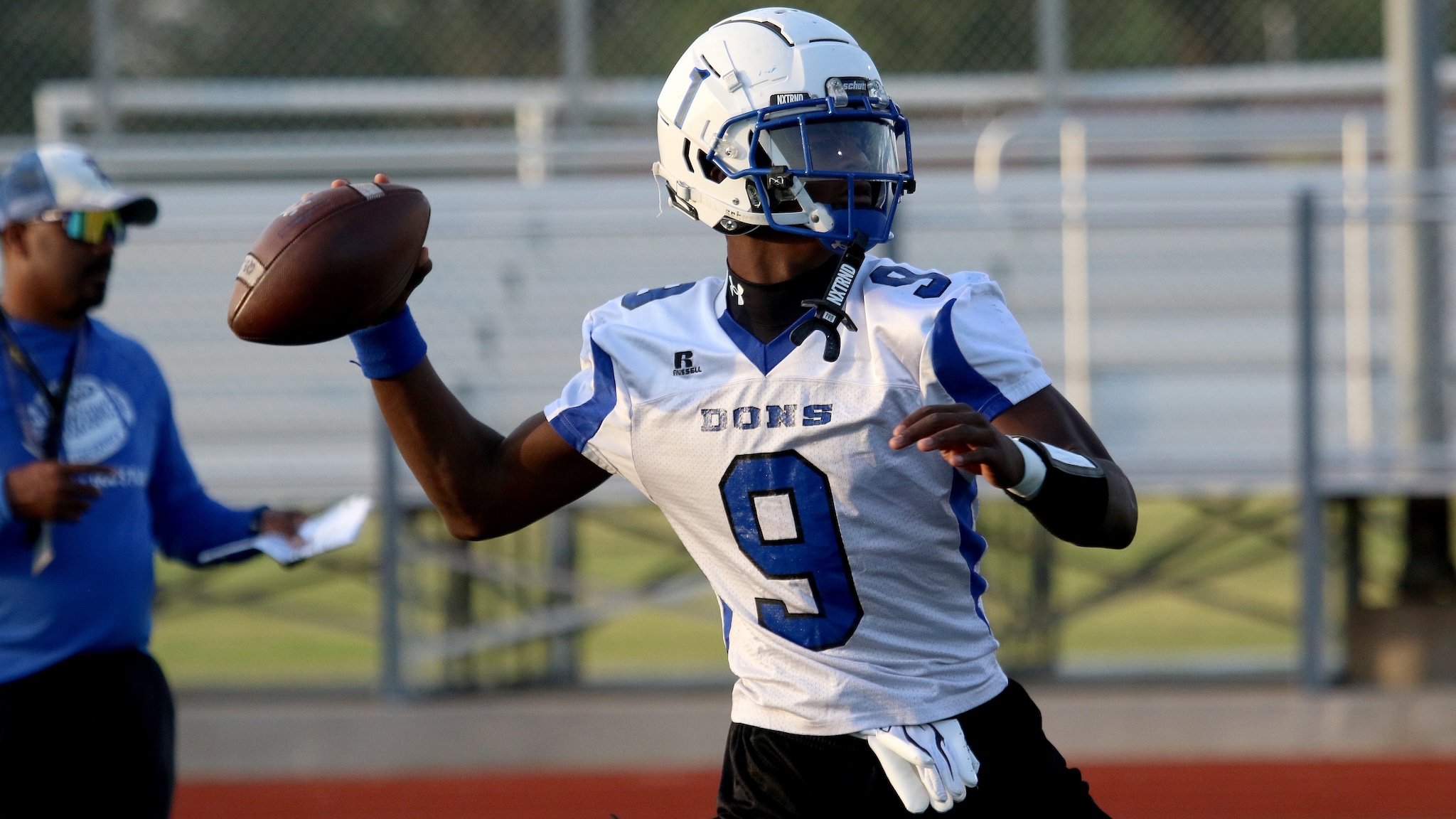 Football player in a white and blue uniform runs with the football, number 9, on a field while a teammate watches from the left.