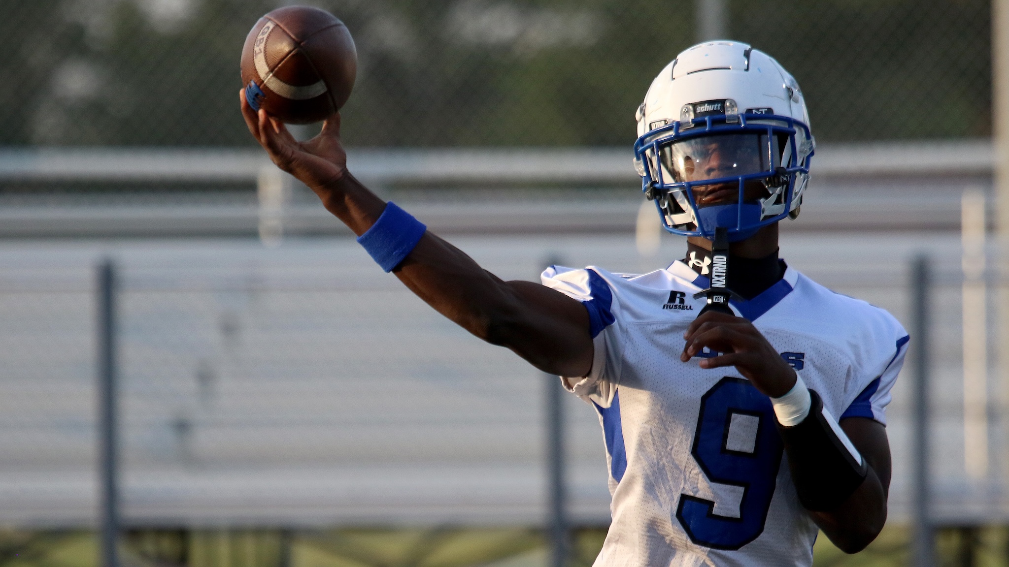Quarterback in a white and blue uniform throwing a football during a game on a fence-lined field.