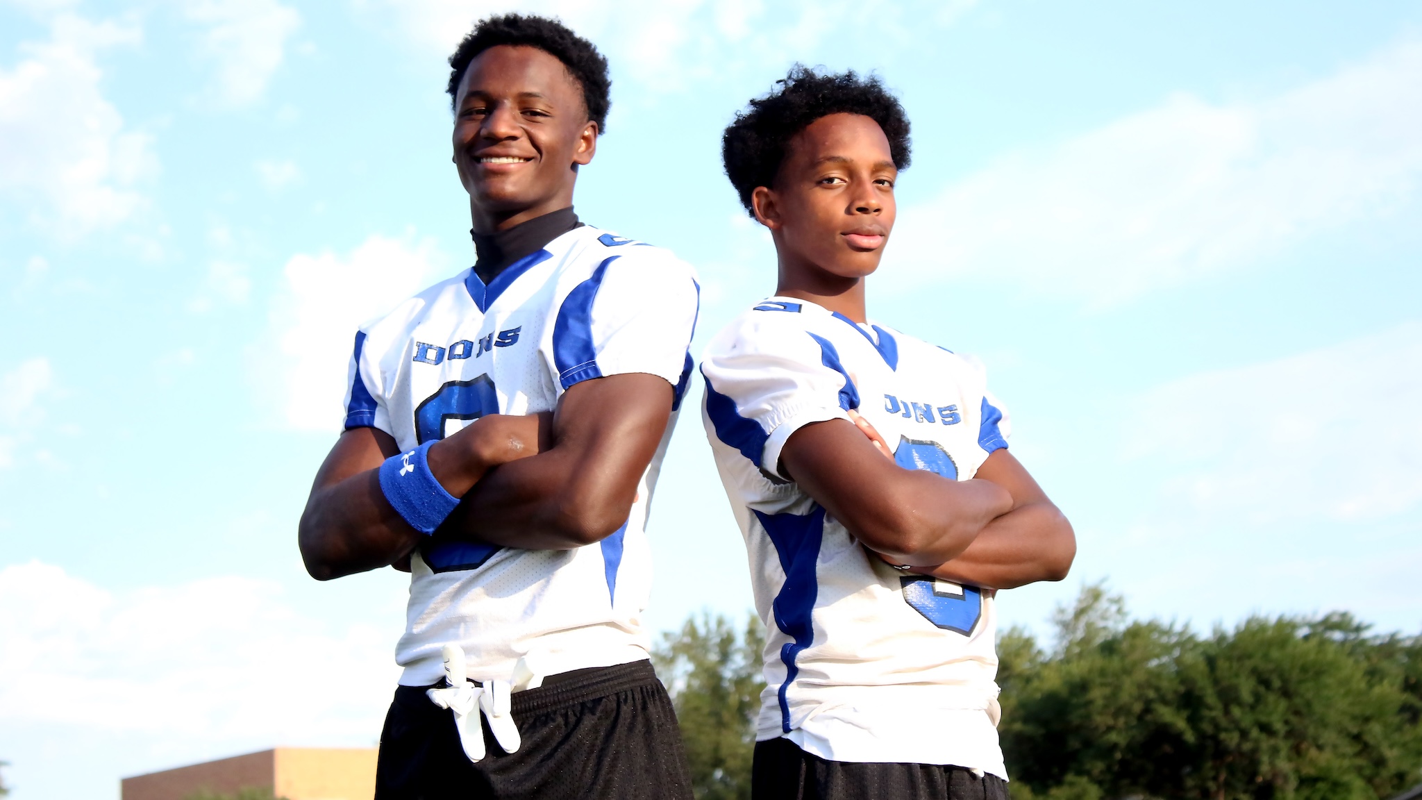 Two young football players pose back-to-back with arms crossed, wearing white and blue uniforms against a clear sky.