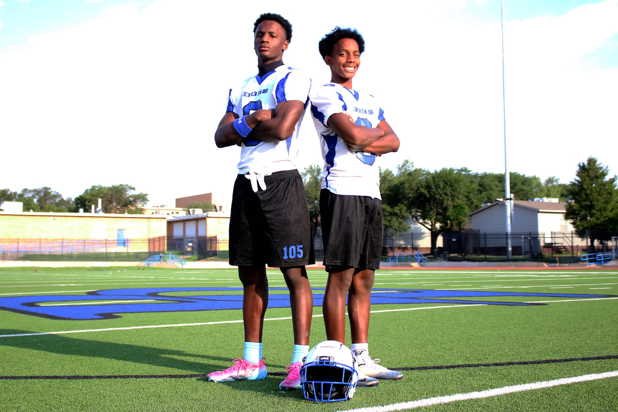 Two male football players in blue-and-white jerseys stand back-to-back with arms crossed on a football field; helmets on the turf at their feet.