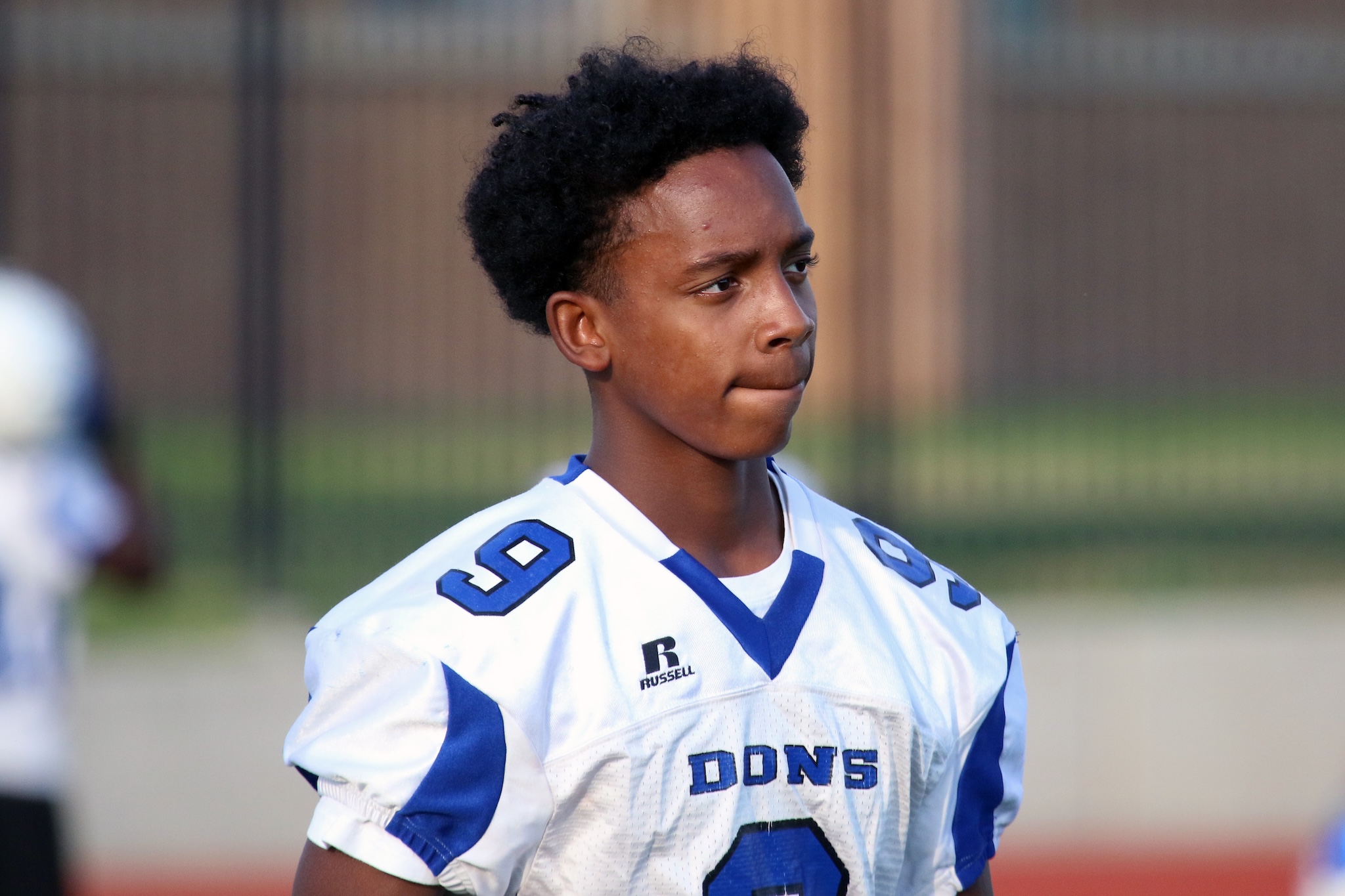 Teenage football player in a white and blue uniform, looking to the right on a field.