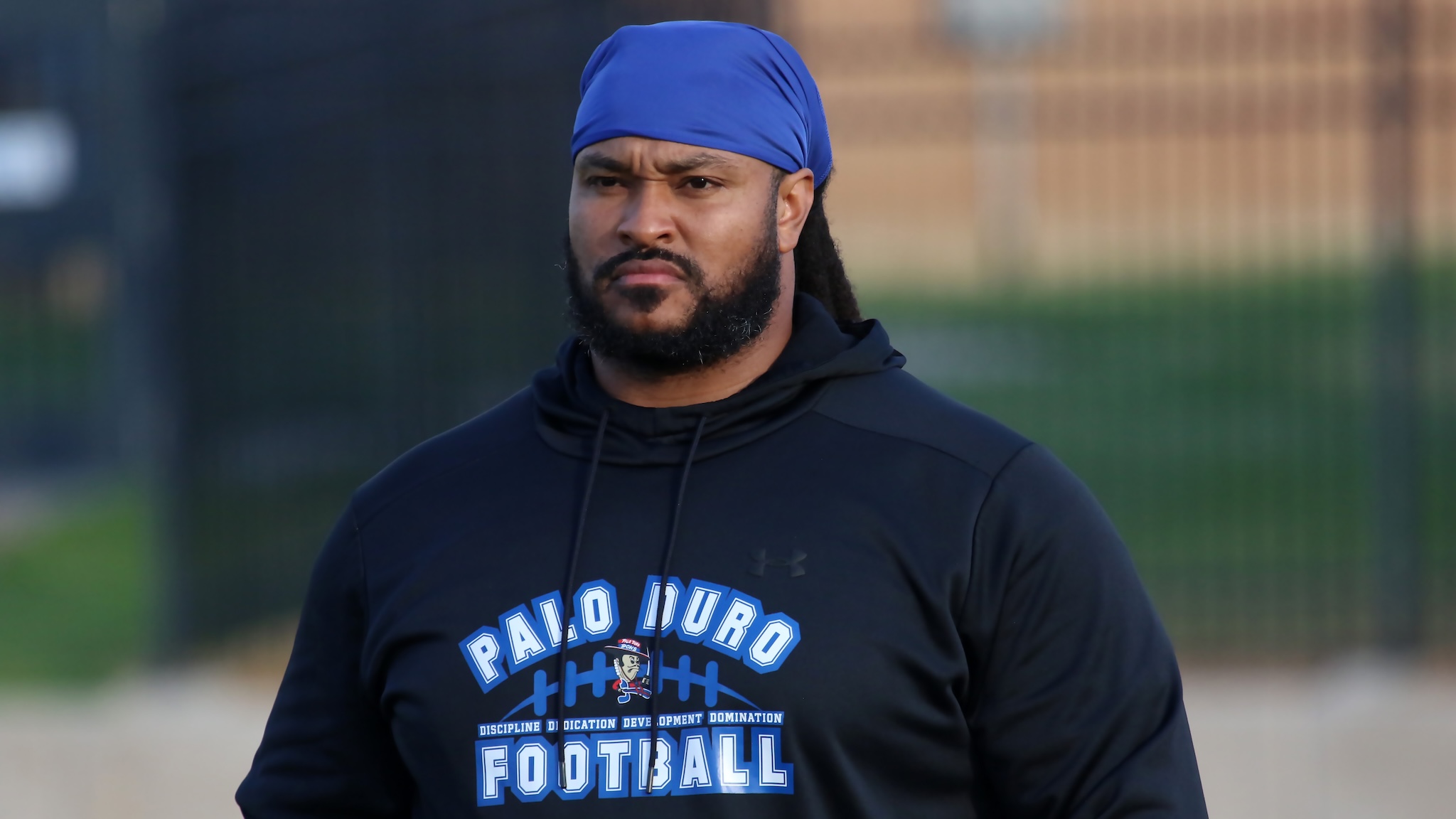 Man wearing a blue head wrap and black hoodie stands outdoors, looking slightly to the side with a serious expression; football graphic on shirt visible