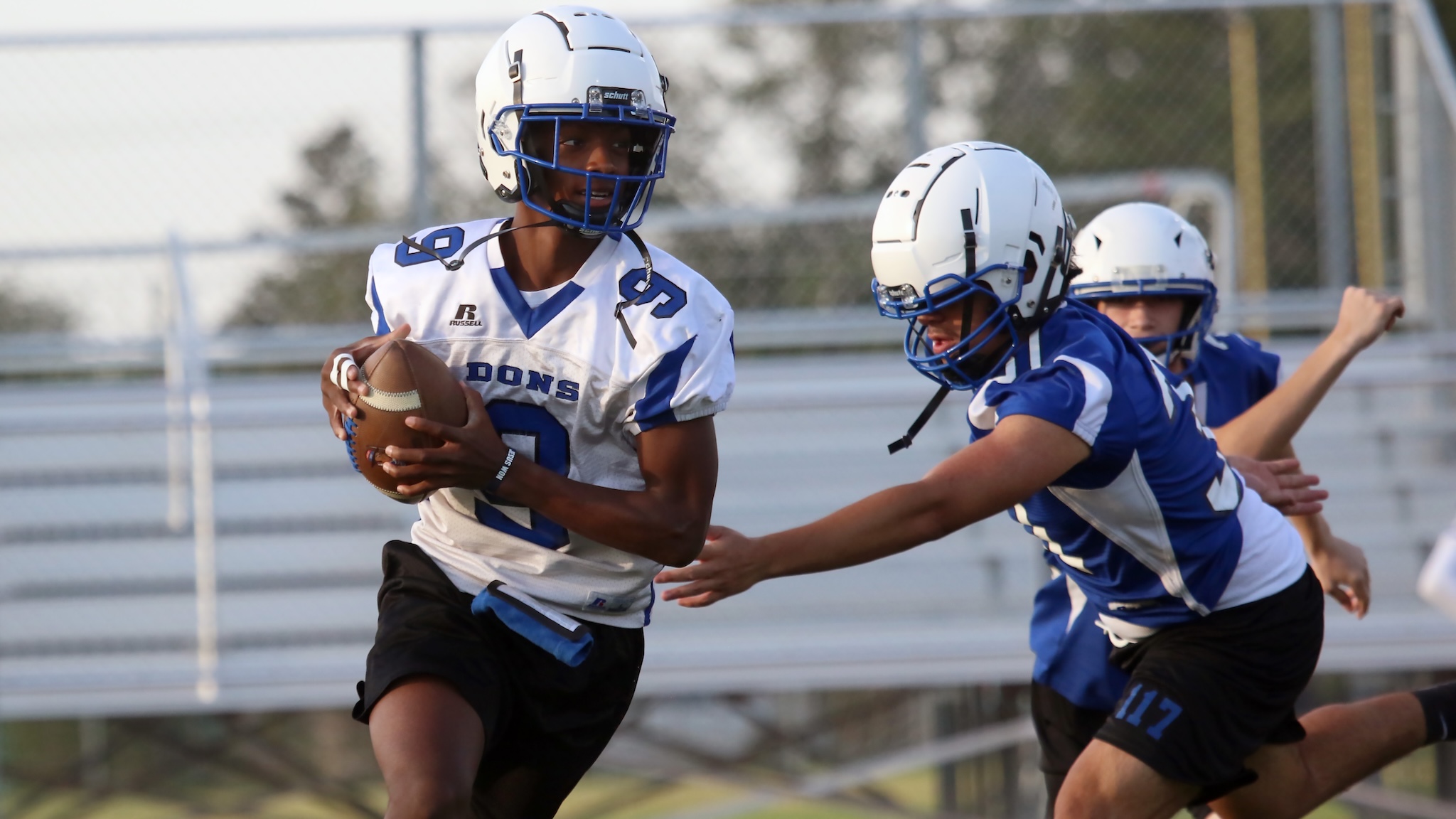 Football player in a white jersey runs with the ball, pursued by blue defenders during a game.