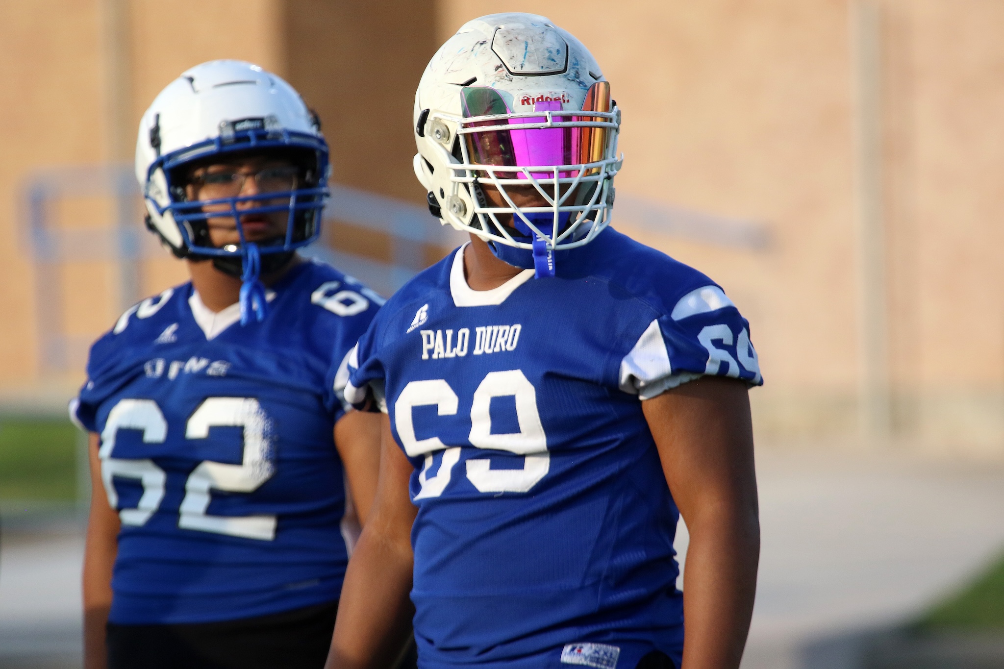Two football players in blue uniforms standing on a field; foreground player wears a white helmet with a pink visor and number 60, Palo Duro on the jersey.