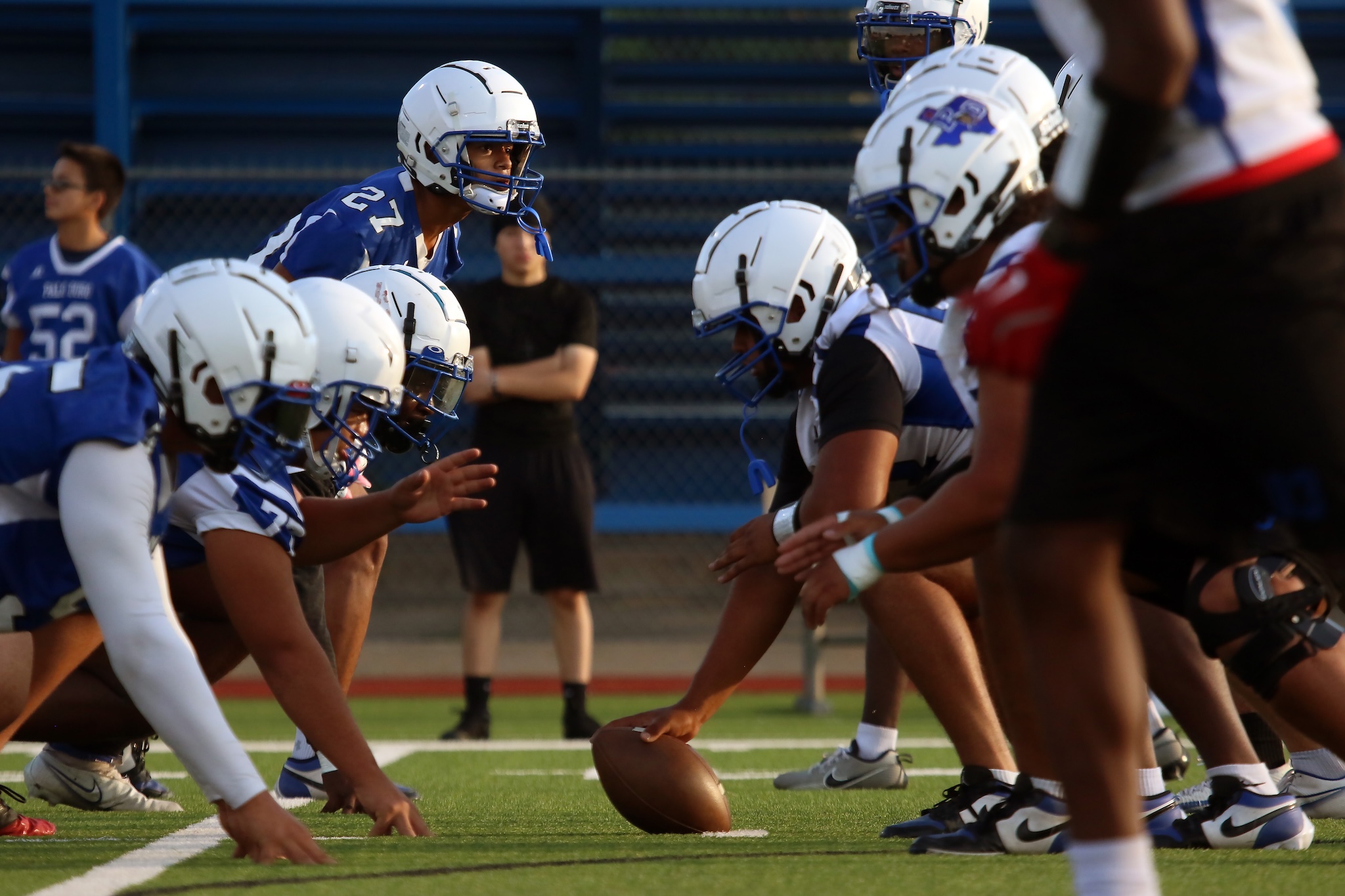 Youth football players in blue and white line up at the line of scrimmage with the ball on the turf, ready for the snap.