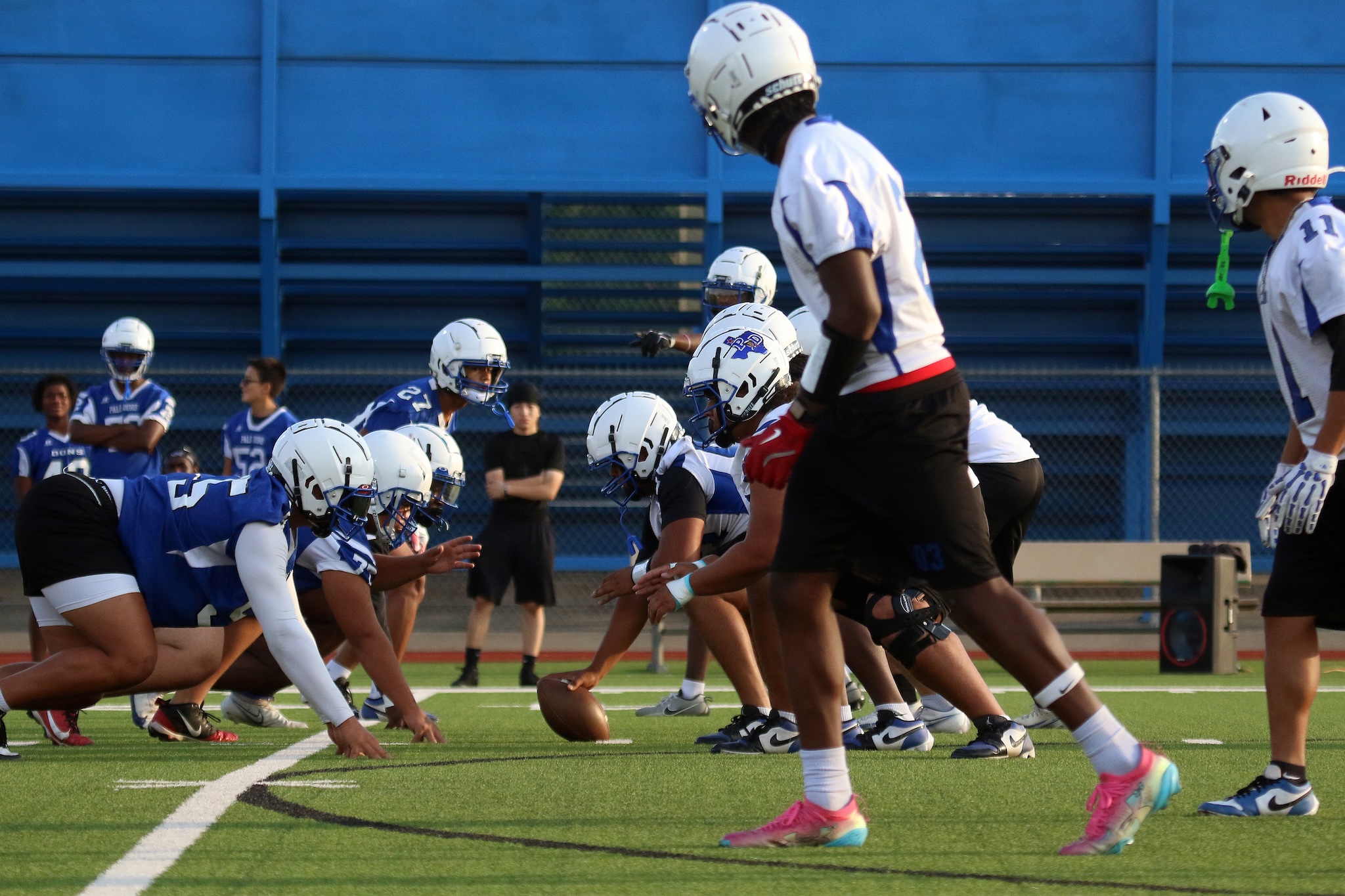 Football players in white and blue uniforms line up at the scrimmage on a green field, ball on the ground ready for a snap.