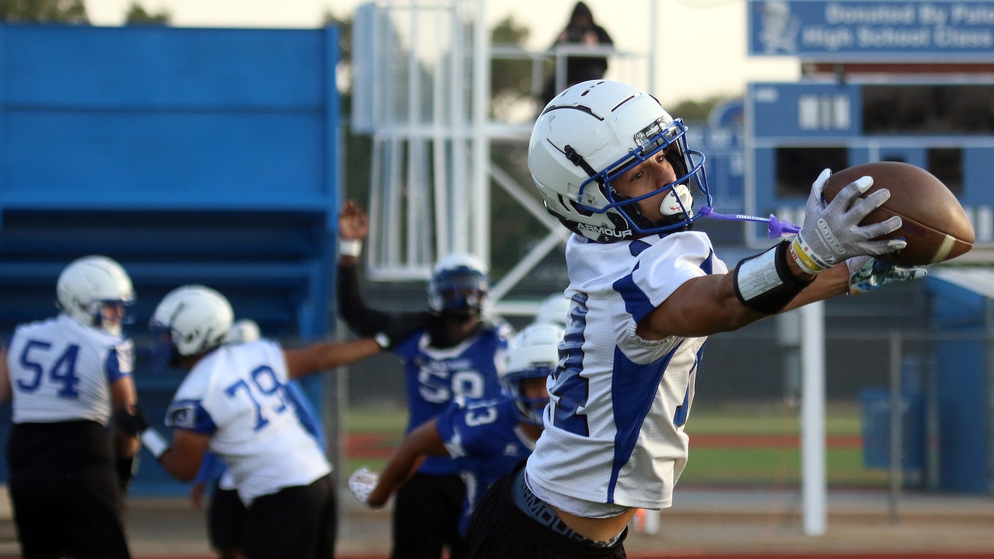 Football player in white and blue uniform reaches for a catch during a game, a blurred stadium in the background.
