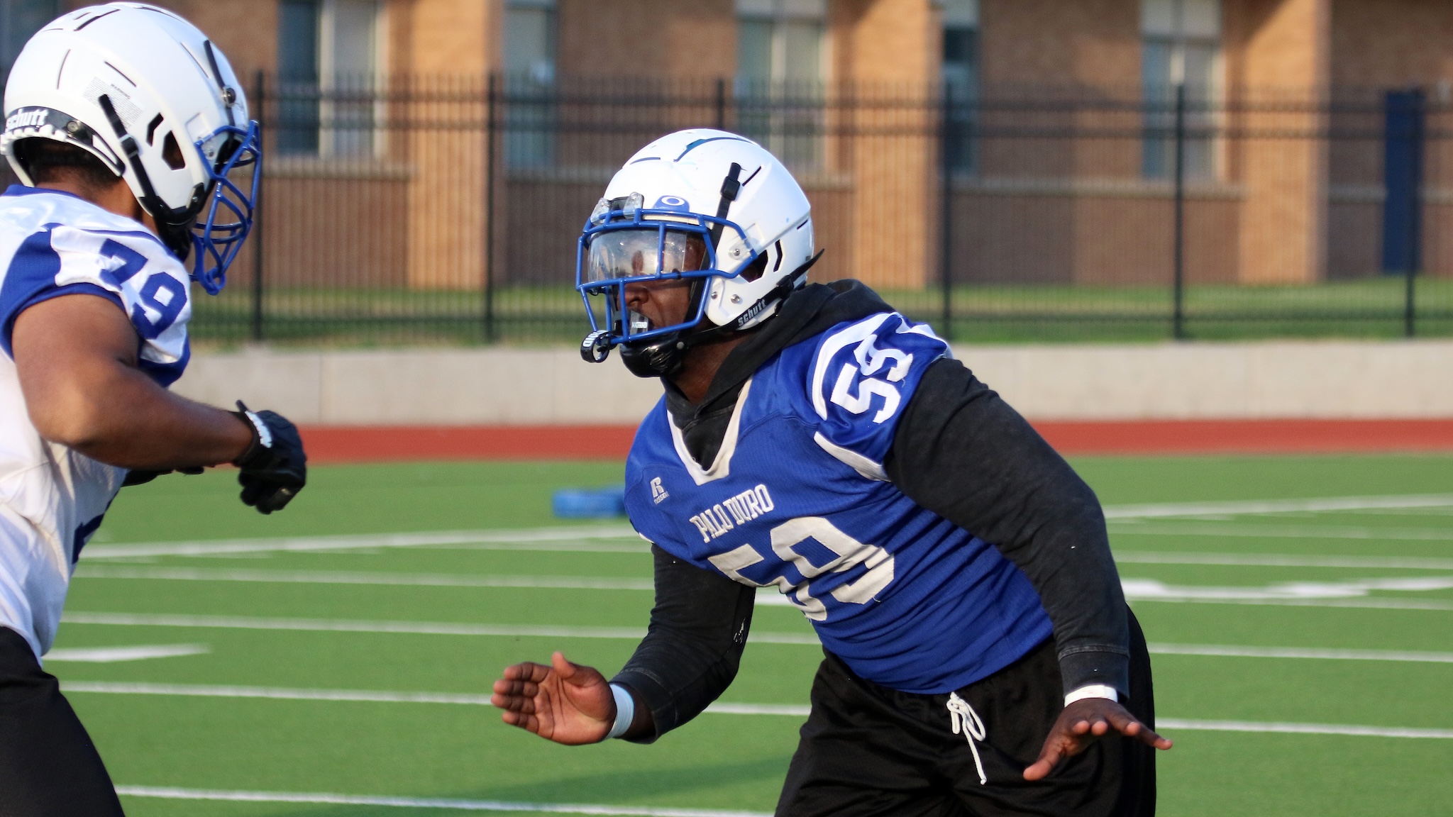 Two football players in blue and white jerseys rush toward each other on a green turf field, helmets on, mid-play action.