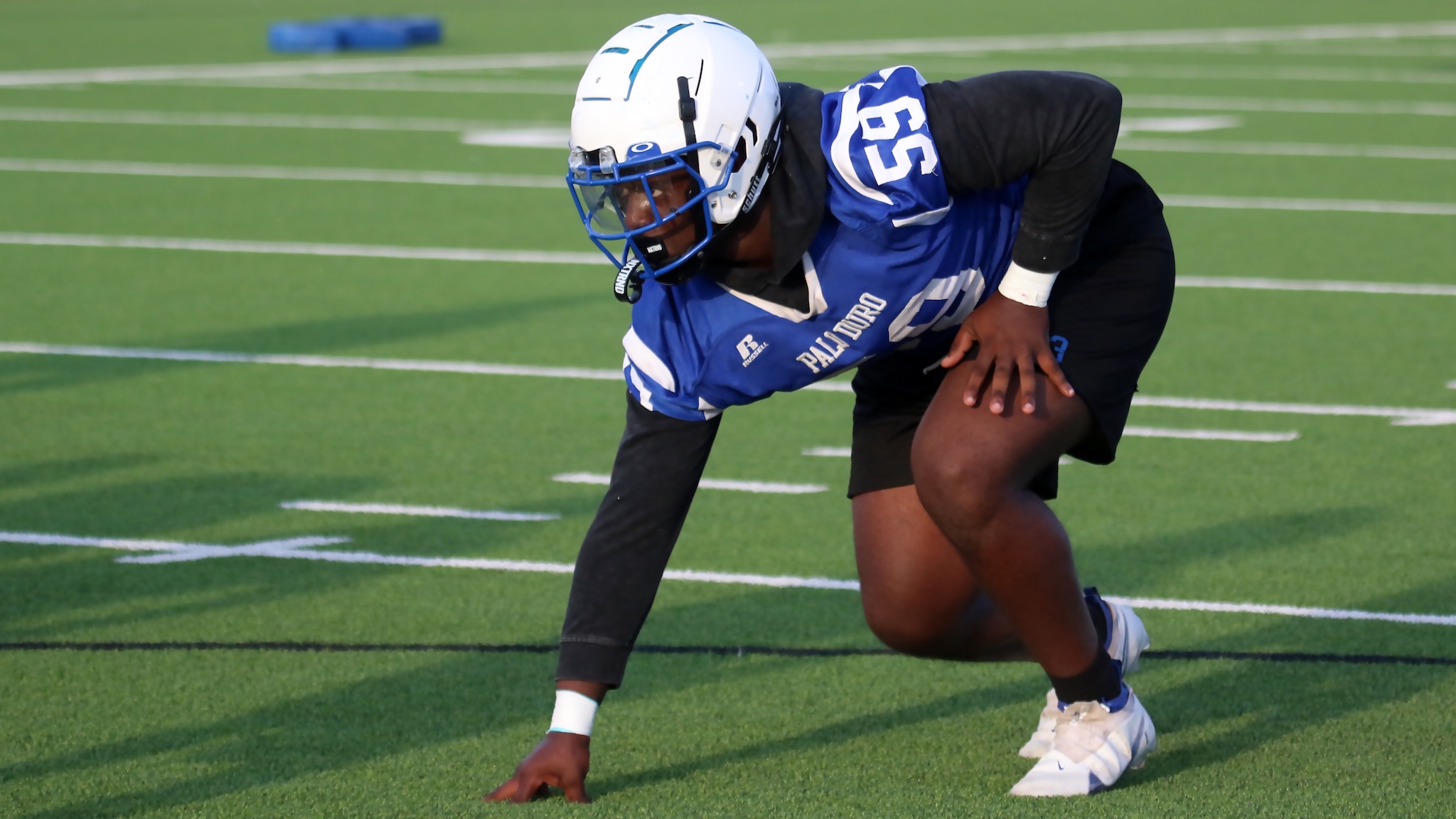 Football player in blue jersey crouching on a green field, helmeted and ready to snap the ball from a low stance.