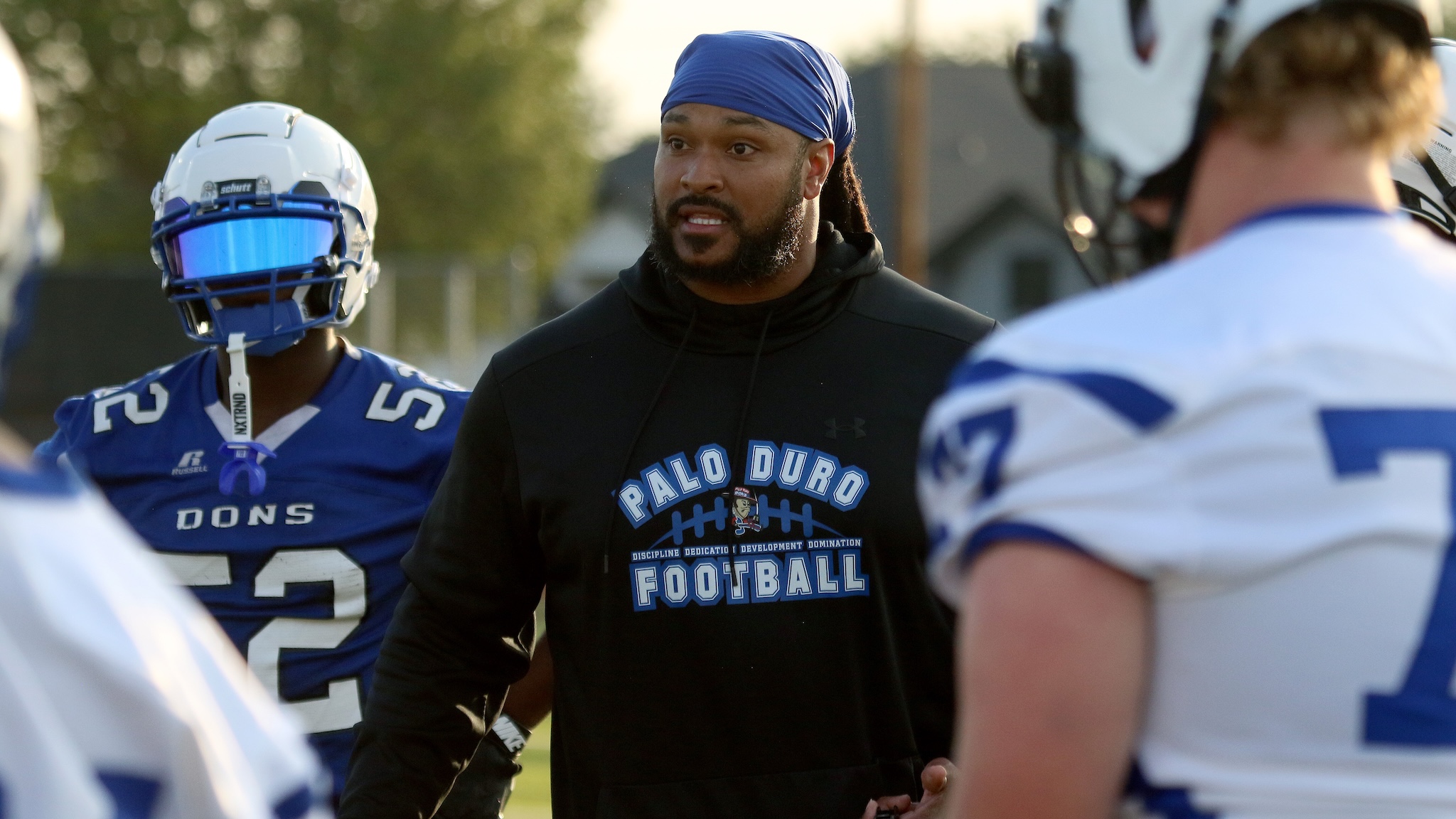 Coach wearing a blue head wrap speaks to players in blue-and-white uniforms during a drill on the field (Palo Duro Football visible on his shirt).