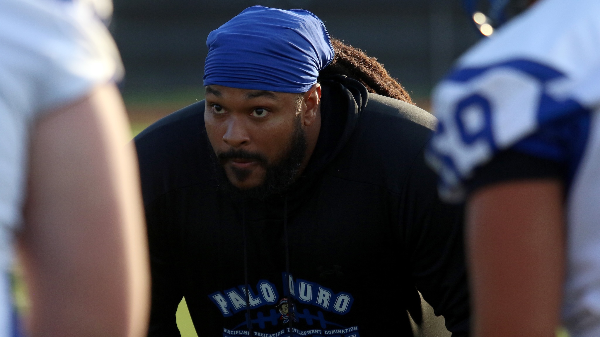 Man with a blue durag and black hoodie leaning forward, focused on something off-camera during a sports practice.