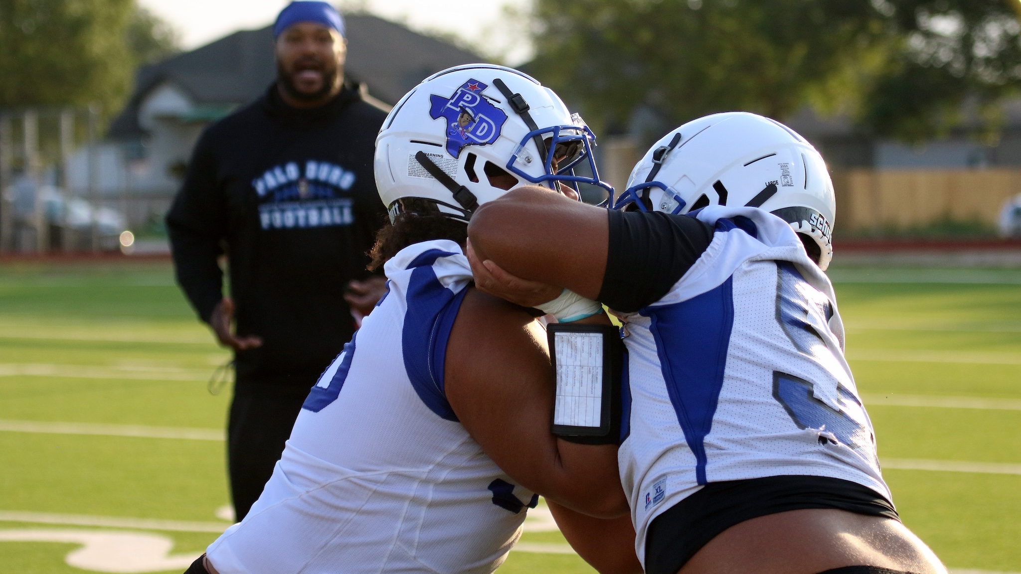 Two football players in white and blue uniforms hug in celebration on the field, coach watching in the background.