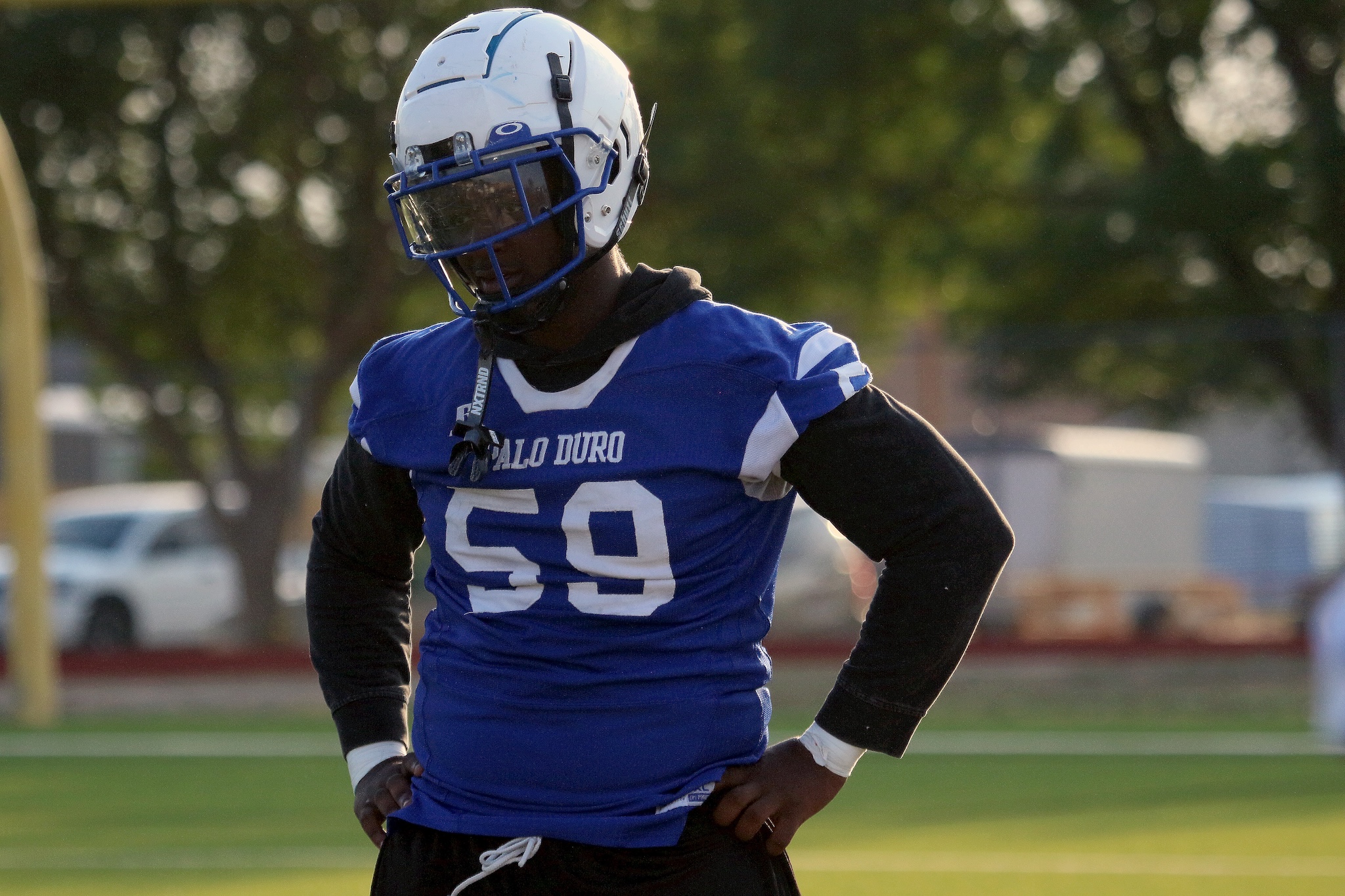 American football player in a blue jersey #59 with a white helmet, standing with hands on hips on a grassy field