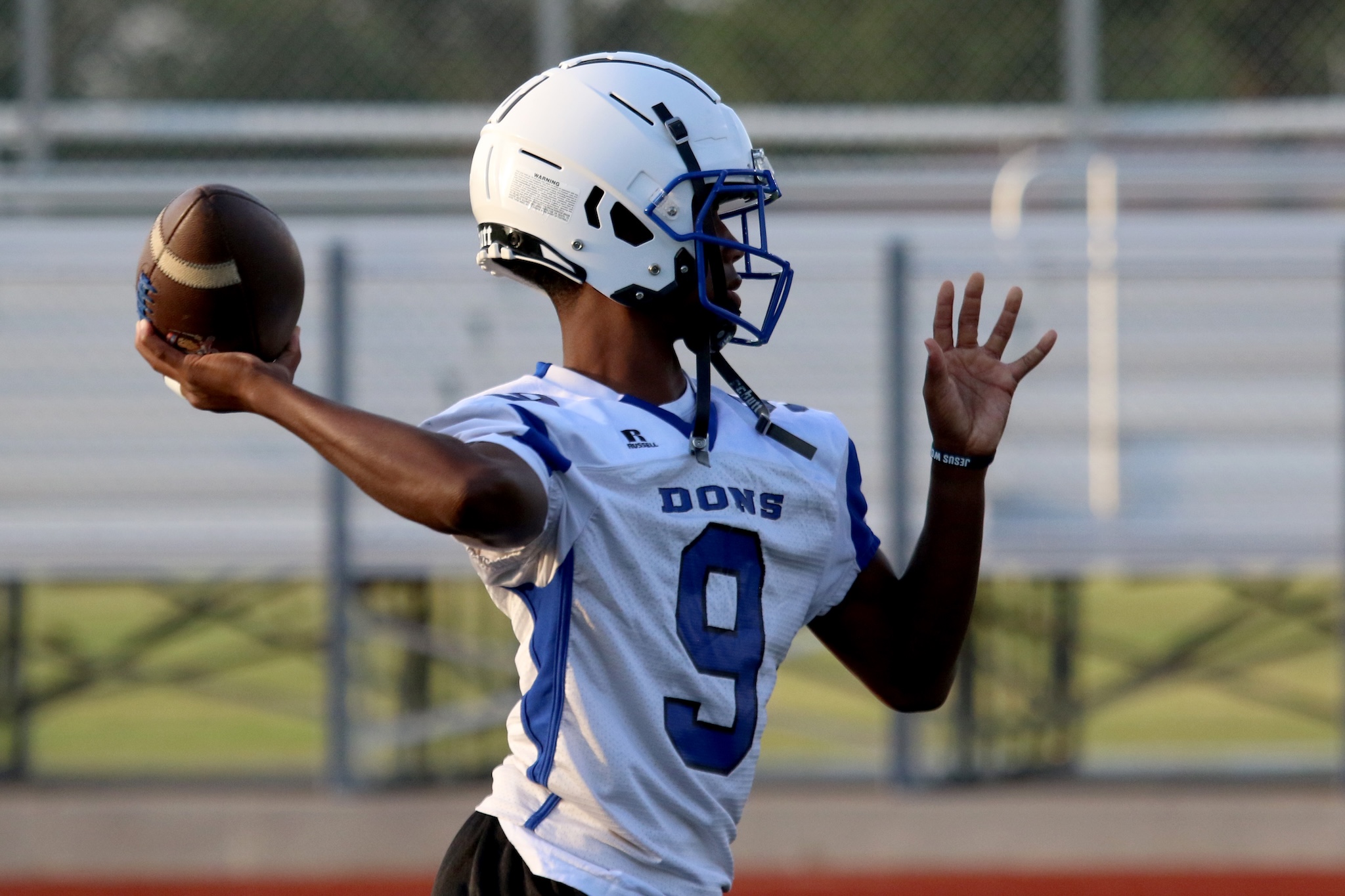 Quarterback in white helmet and jersey throwing a football on a field with chain-link fence in the background.