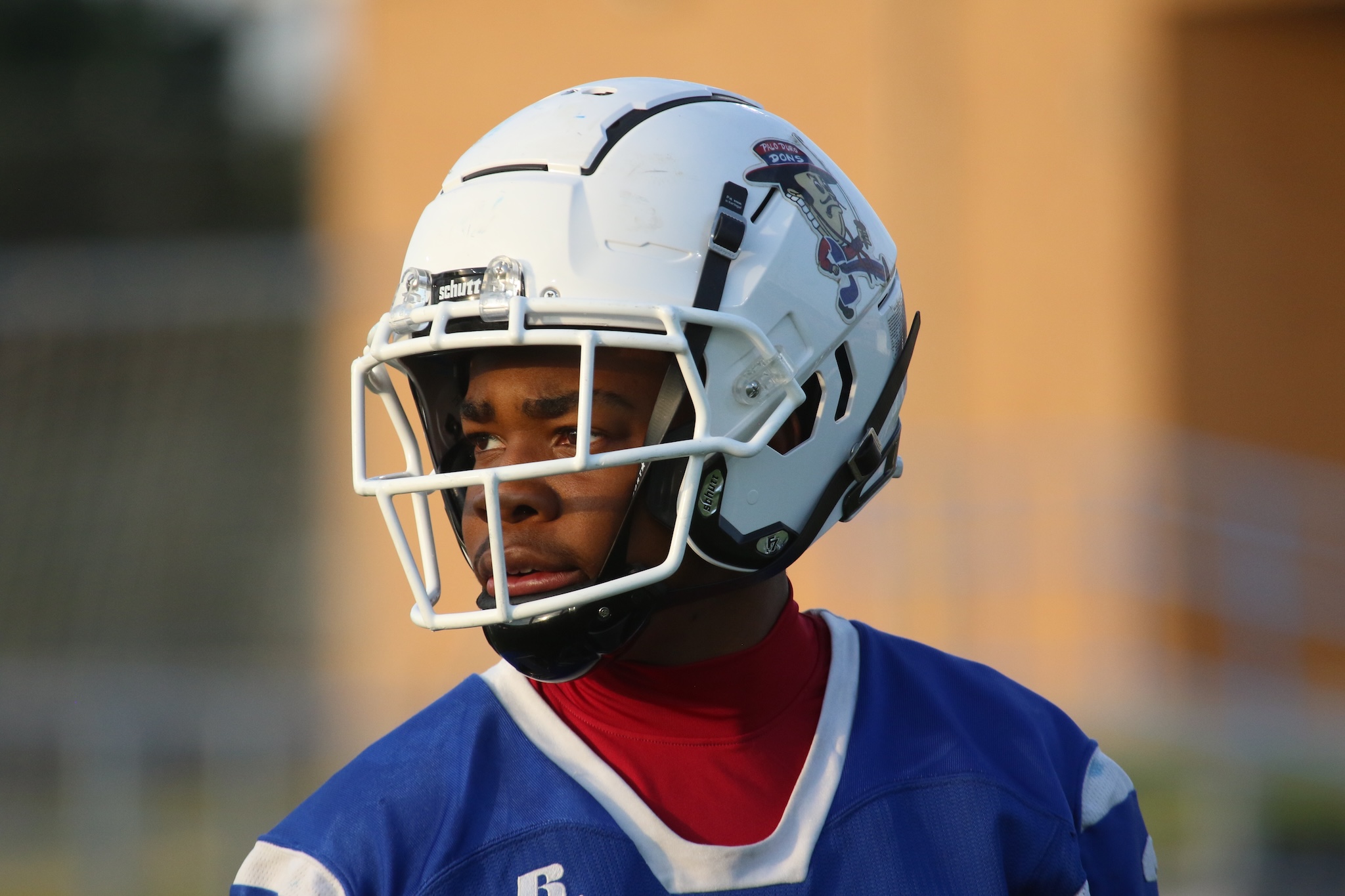 Close-up of a young football player wearing a white helmet with a faceguard and a blue jersey, looking to the left.
