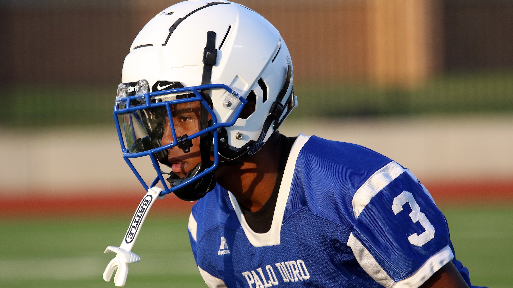 Close-up of a lacrosse player wearing a white helmet with blue face cage and a blue jersey on a sunny field.