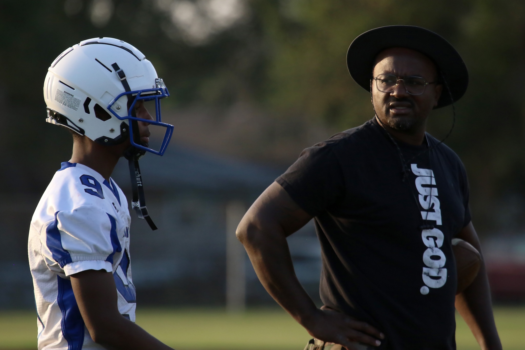 Football player in a white and blue helmet talks with a coach wearing a black shirt and wide-brim hat on a field.
