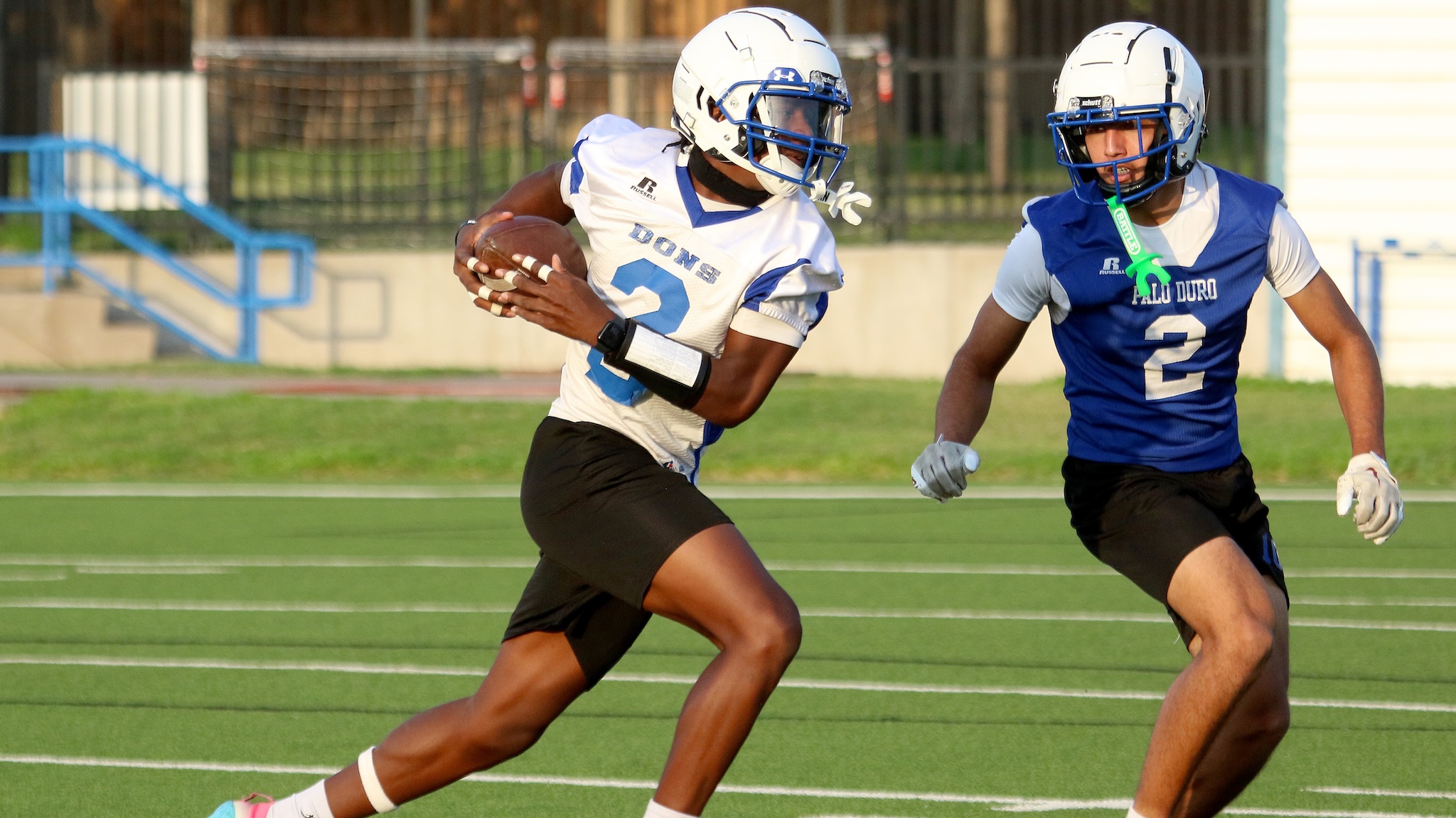 Two football players sprint on a green field; the ball carrier wearing a white jersey and helmet is chased by a defender in blue.