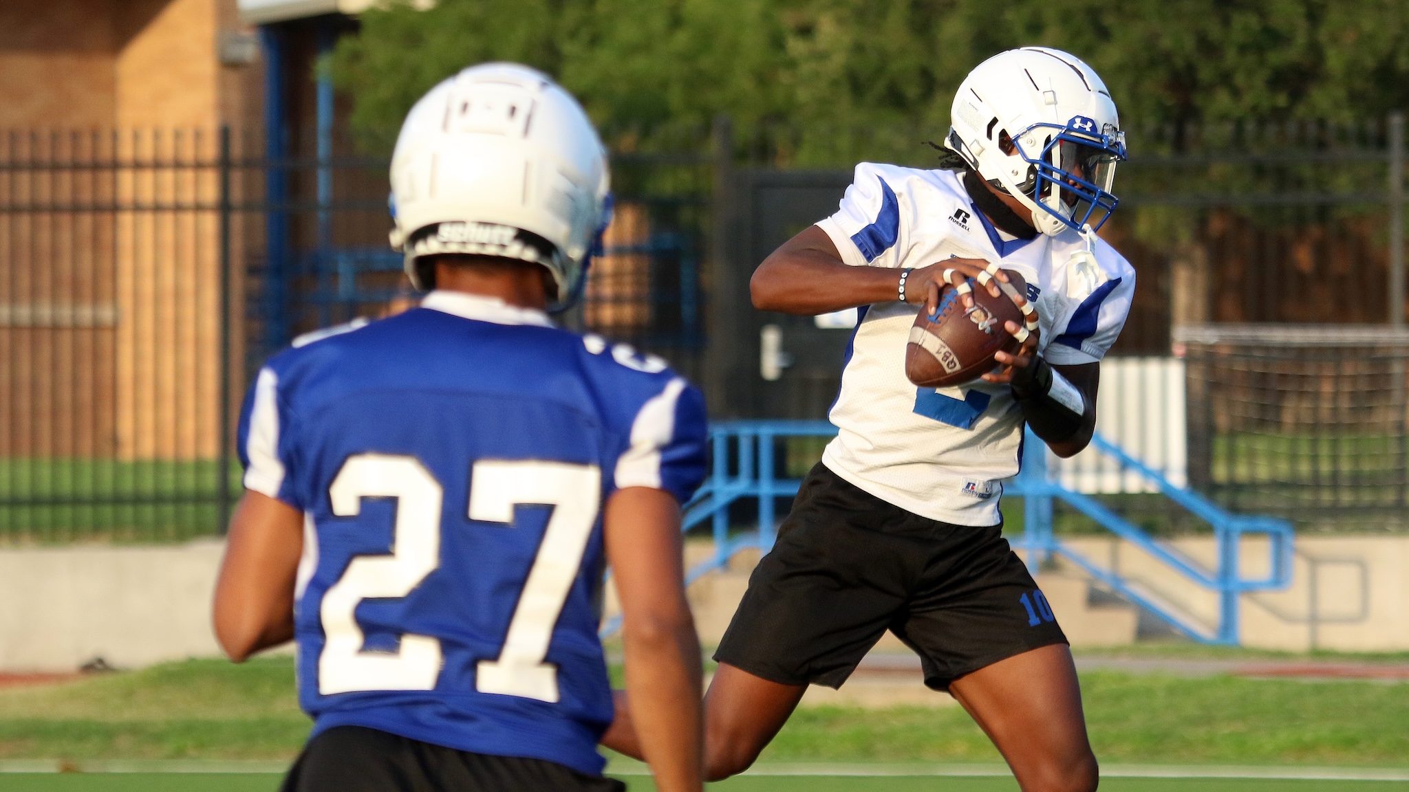 Football player in a white jersey with blue accents cradles the ball as he runs toward the right, pursued by a defender in blue.