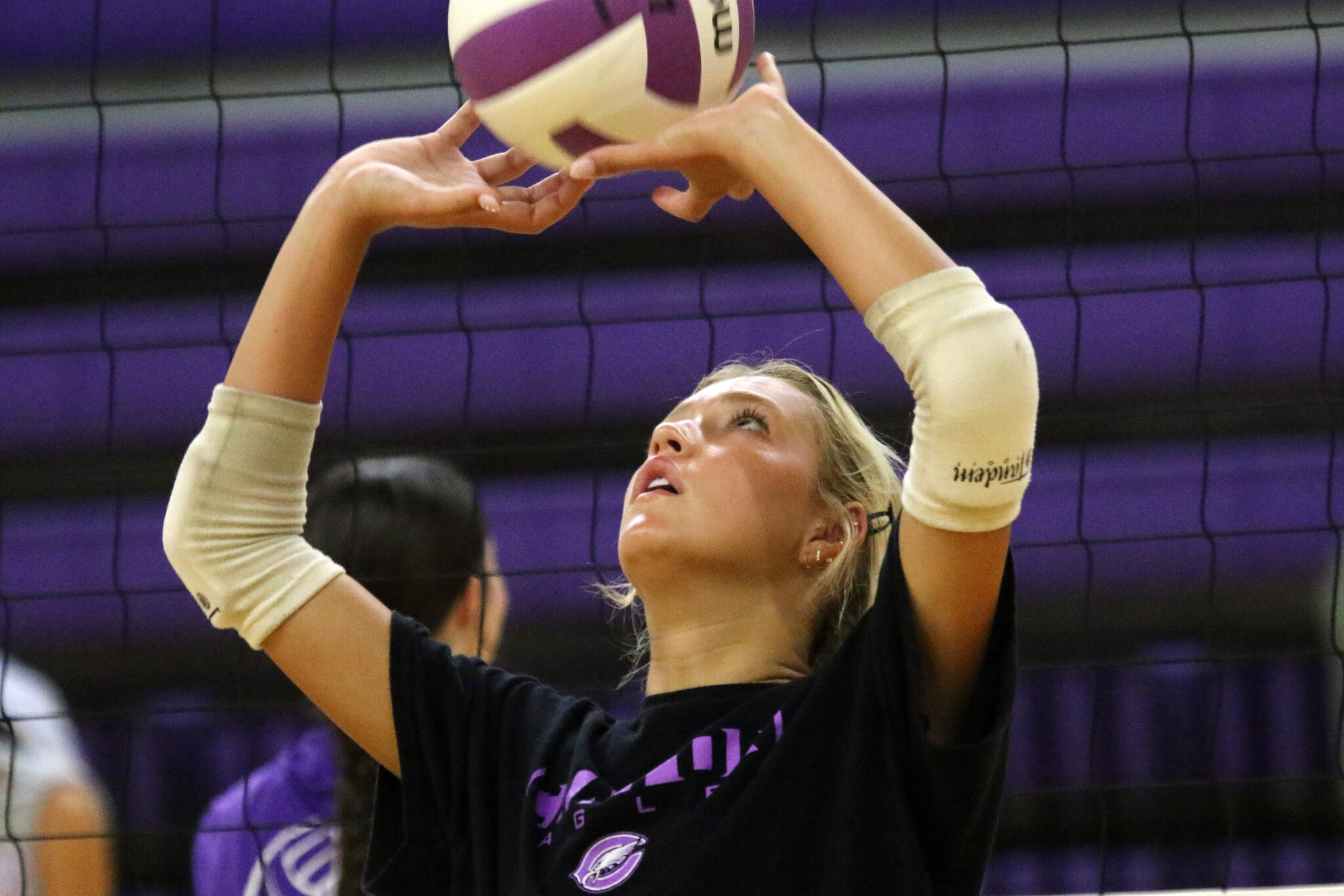 1K6A4422 | Press Pass Sports Female volleyball player with beige arm sleeves reaching up to touch a purple-and-white ball near the net.
