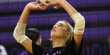 Female volleyball player with beige arm sleeves reaching up to touch a purple-and-white ball near the net.