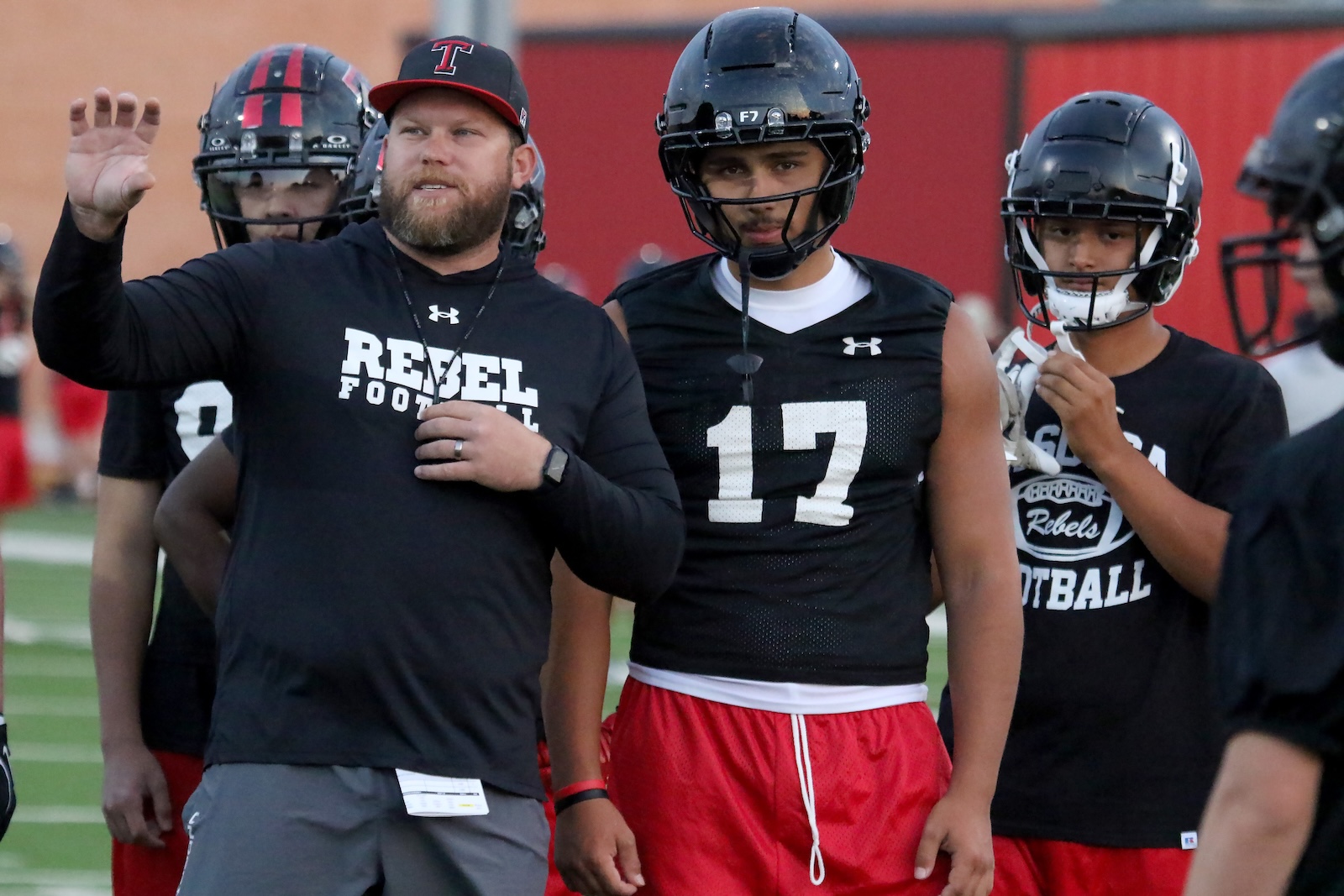 Football coach with a beard waves to players during practice; players in helmets and black Rebels shirts standing on the field