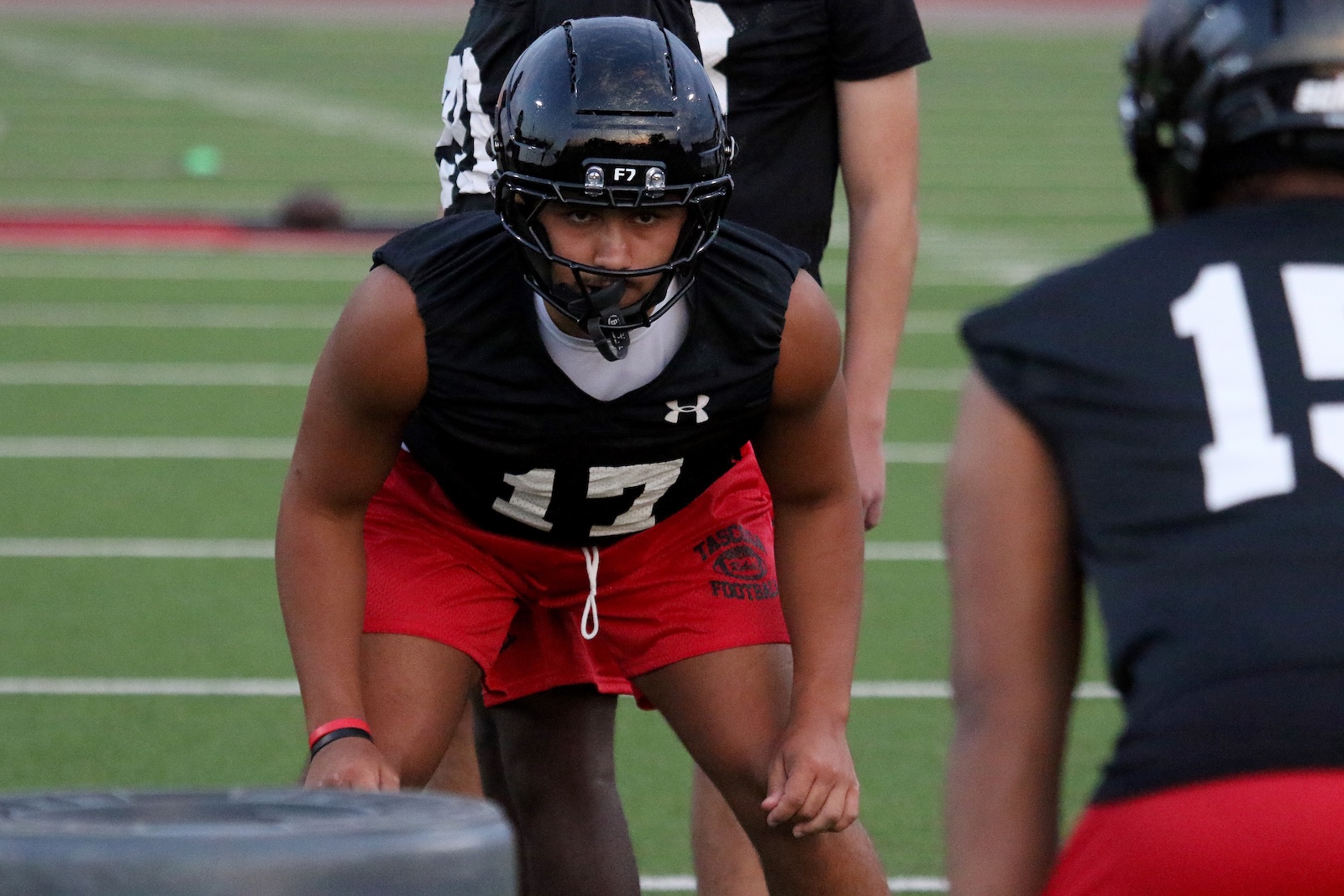 Football player in black jersey (#17) crouches on a turf field, wearing a helmet and red shorts, ready for a drill.