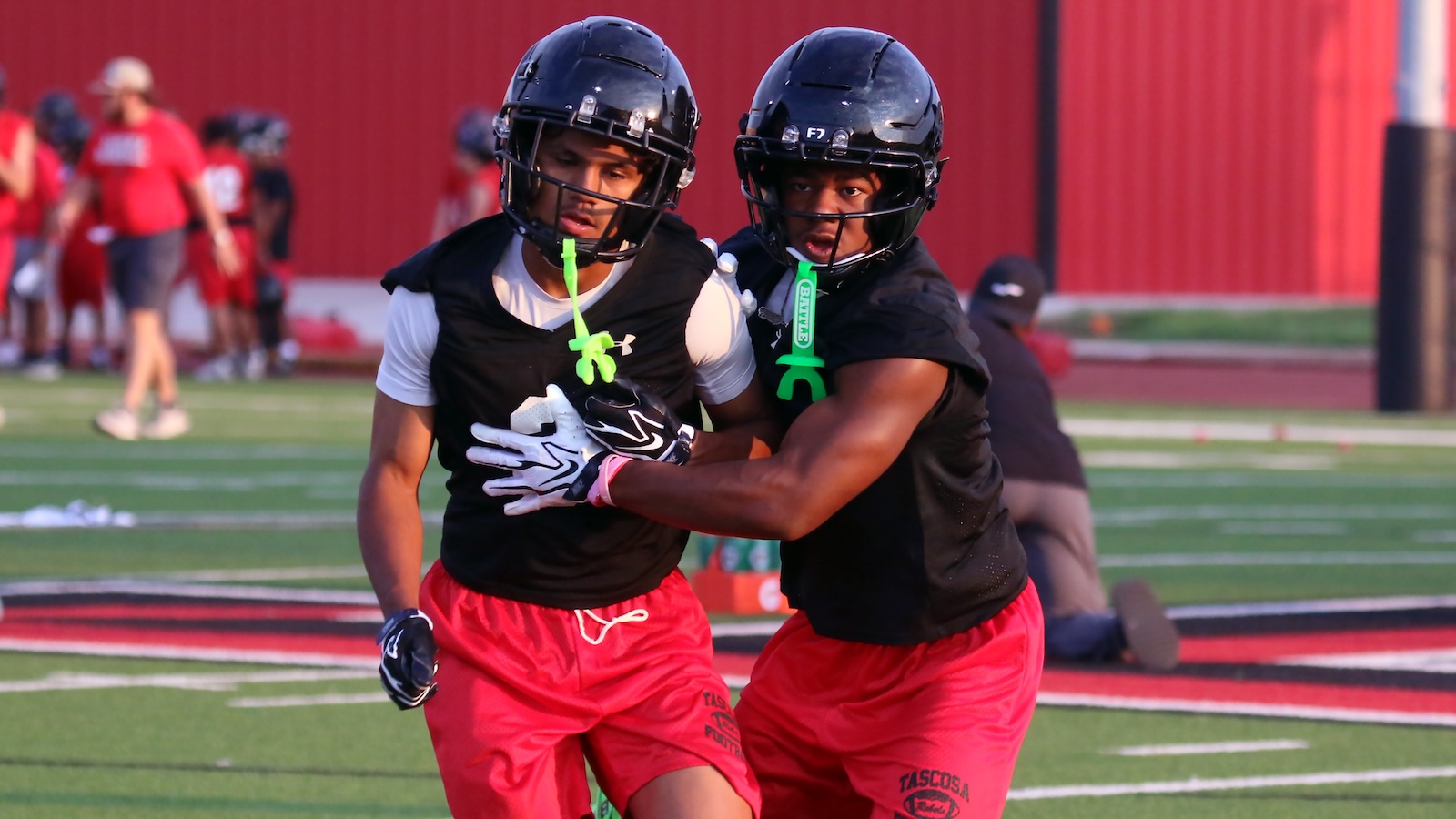Two football players in black jerseys and red shorts tackle during practice on a turf field, wearing helmets and gloves.