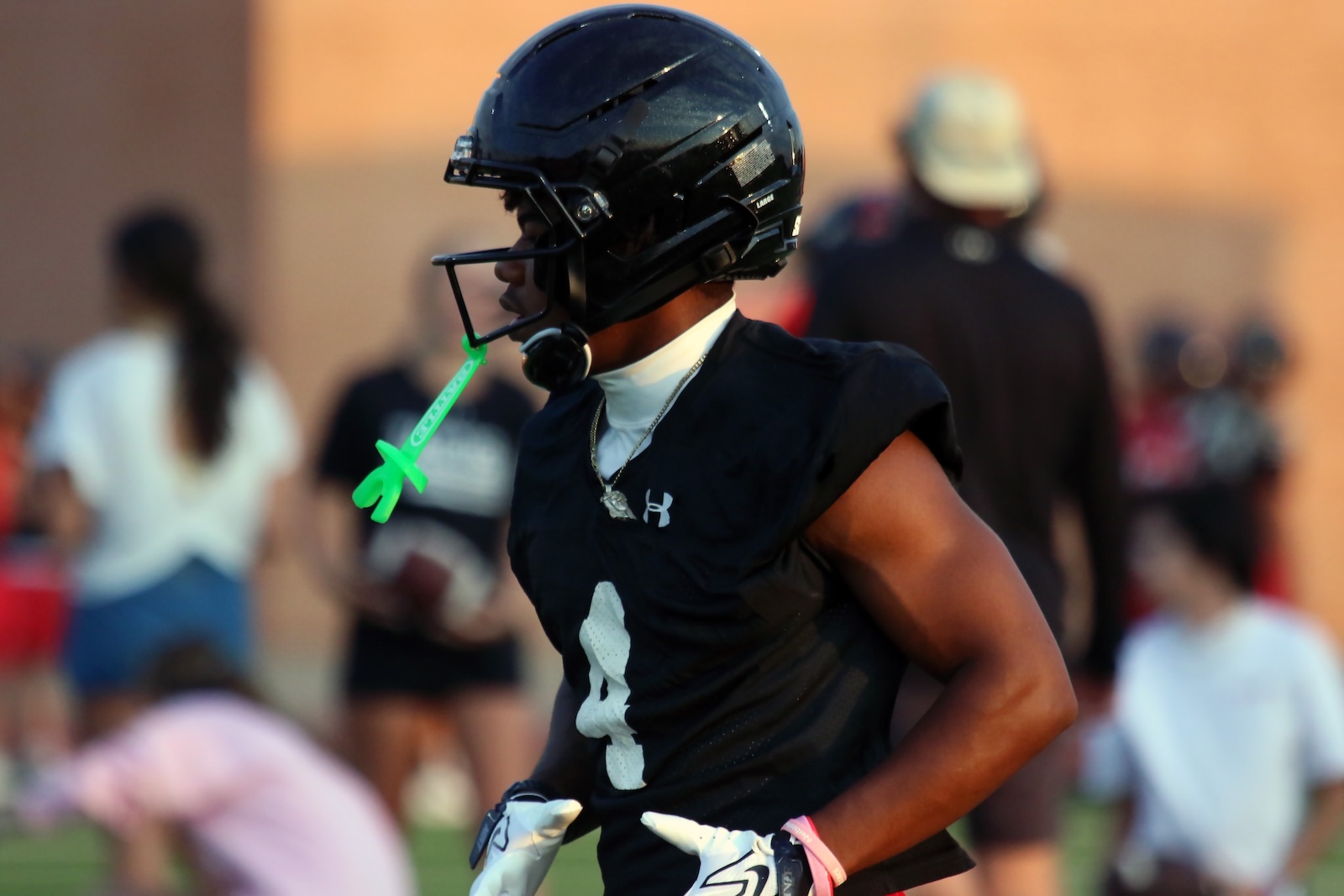 Football player in black practice jersey and helmet, neon green mouthguard attached, on a blurred crowd-filled field.