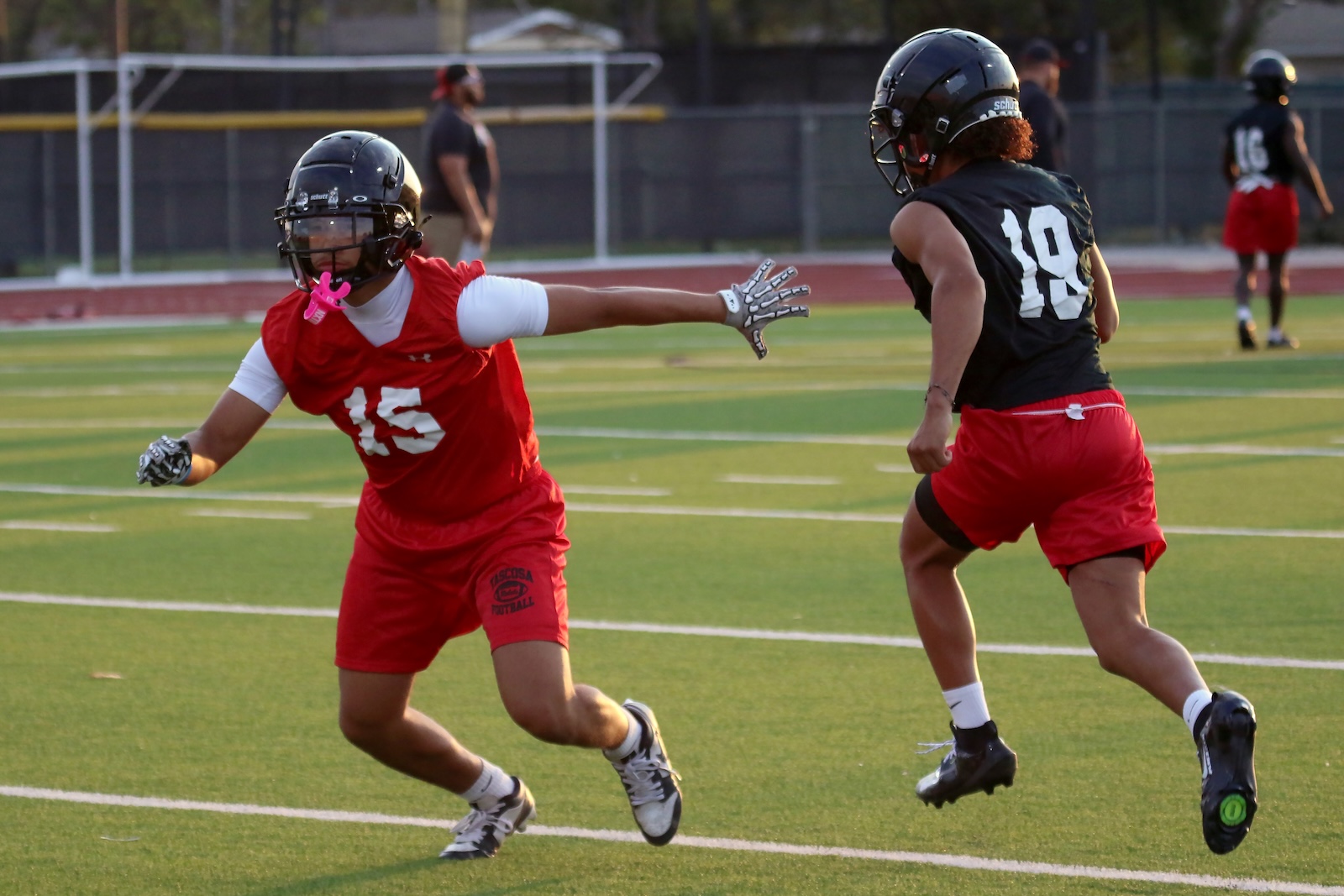 Two football players sprint on a green turf field; the left player wearing red with #15 reaches out as the right player in black #10 runs by.