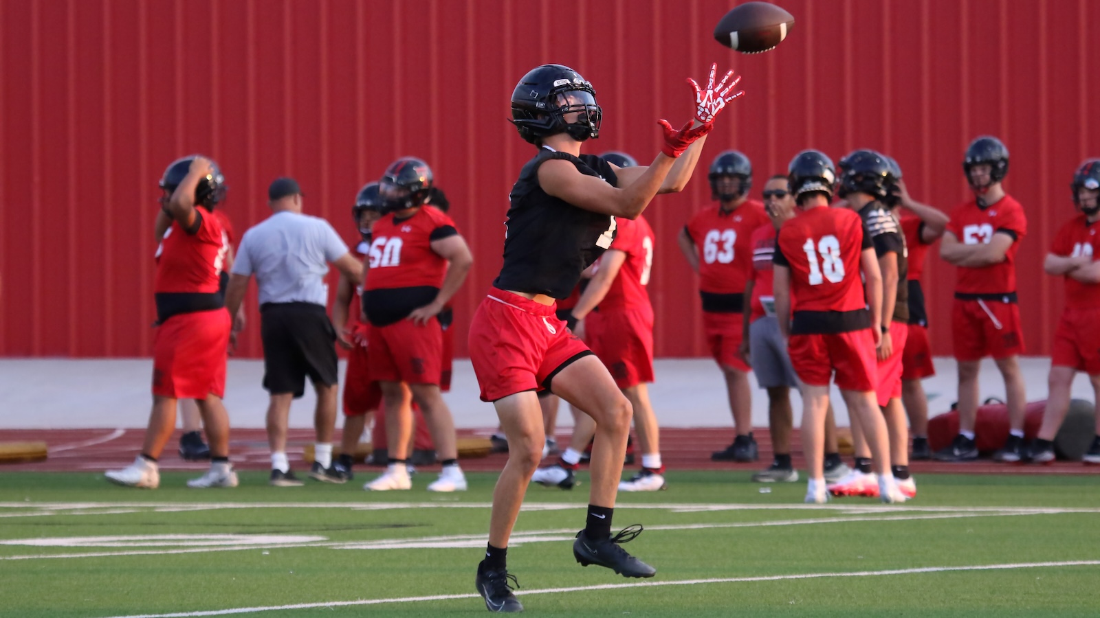 Player in a black jersey and red shorts leaps to catch a football during practice, with teammates in red uniforms watching in the background.
