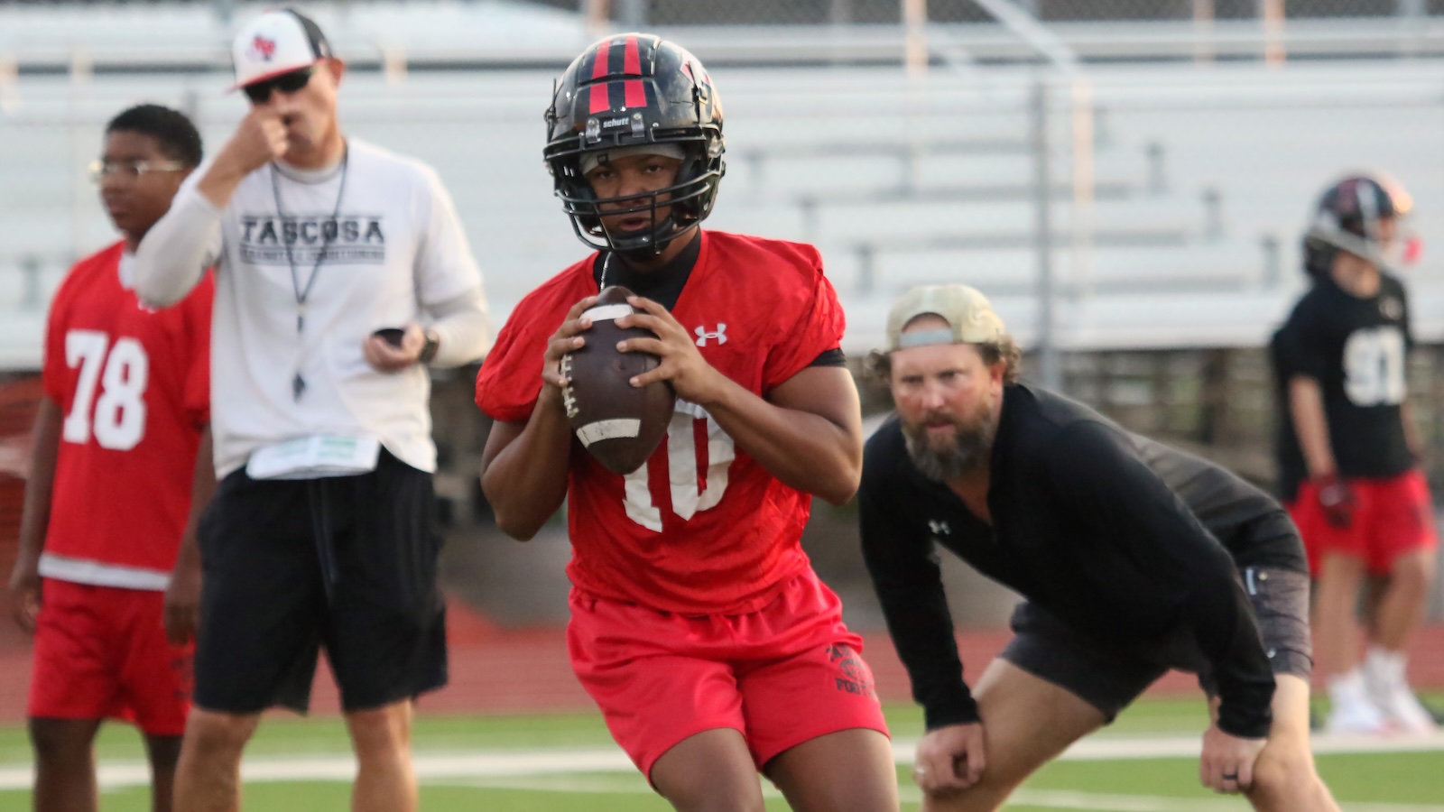Youth football quarterback in red jersey (#10) grips a football, ready to throw/fake on a practice field. Behind him, coach and teammates watch.