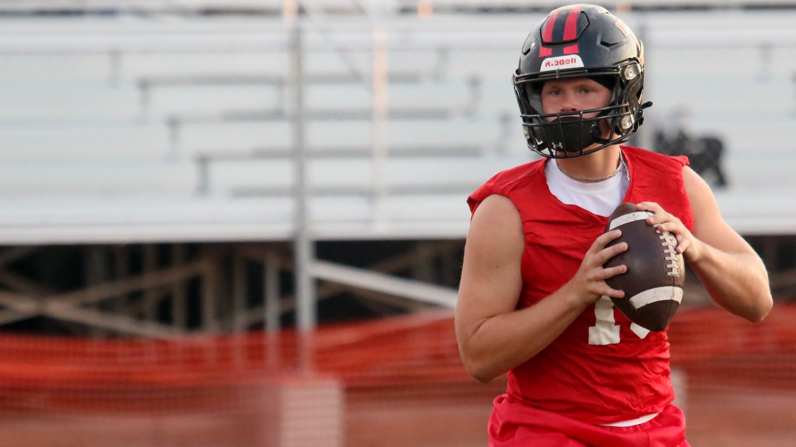 Football quarterback in a red jersey with a black helmet, gripping a football and preparing to pass on the field.