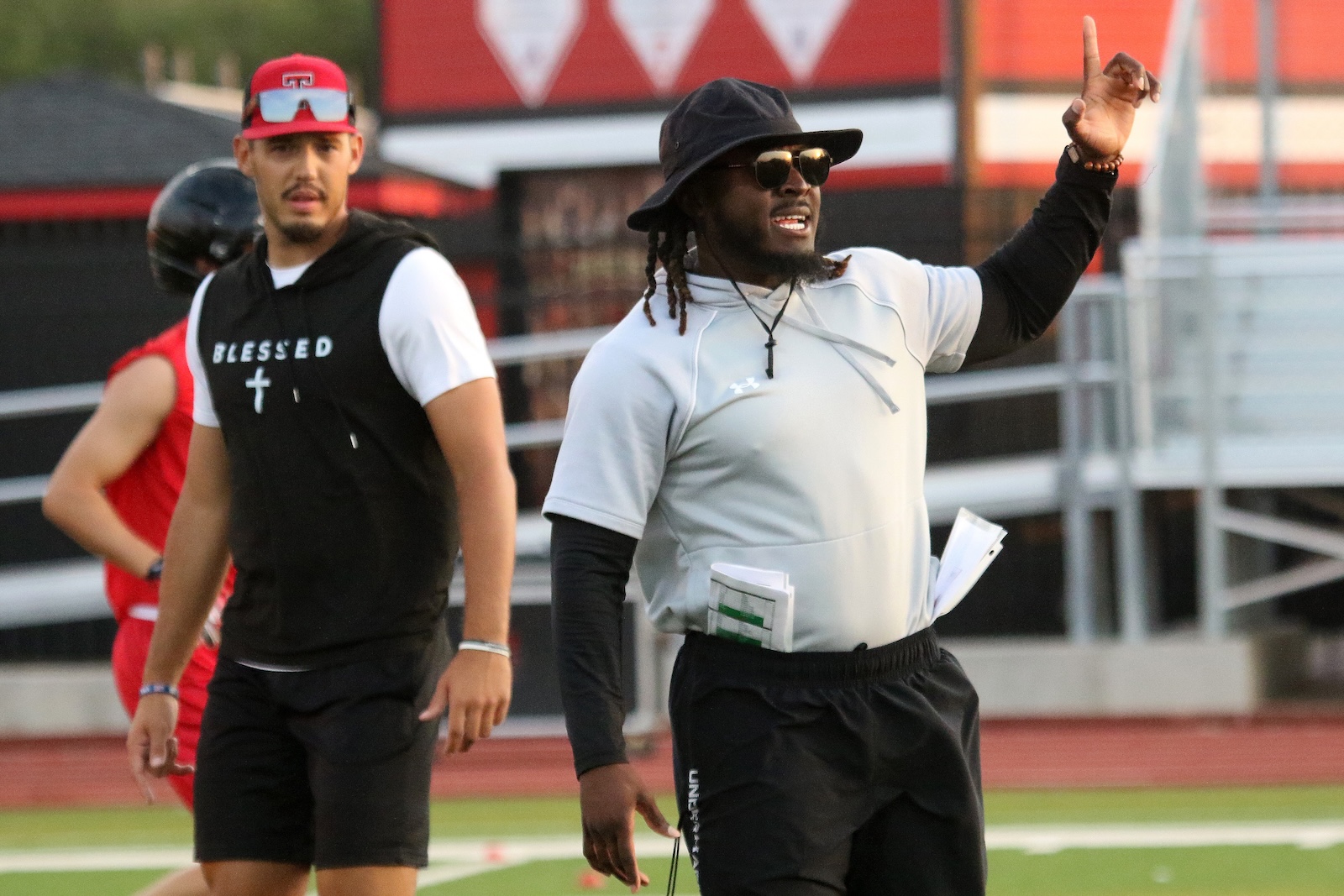 Coach in gray athletic shirt and black bucket hat points upward while speaking on a football field; teammate in a black sleeveless shirt with'BLESSED' chevron stands nearby.