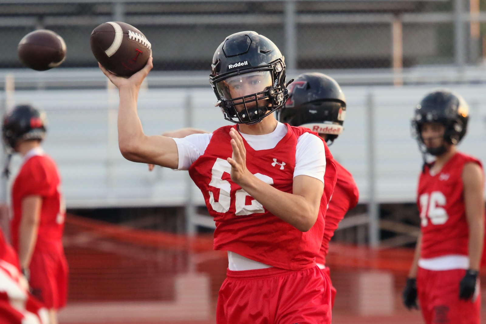 Quarterback in a red jersey and black helmet throwing a football on a practice field with teammates in the background