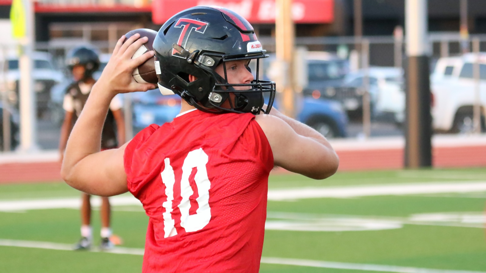 Quarterback in a red jersey with number 10 throwing a football on a green turf field, wearing a black helmet with a red logo.