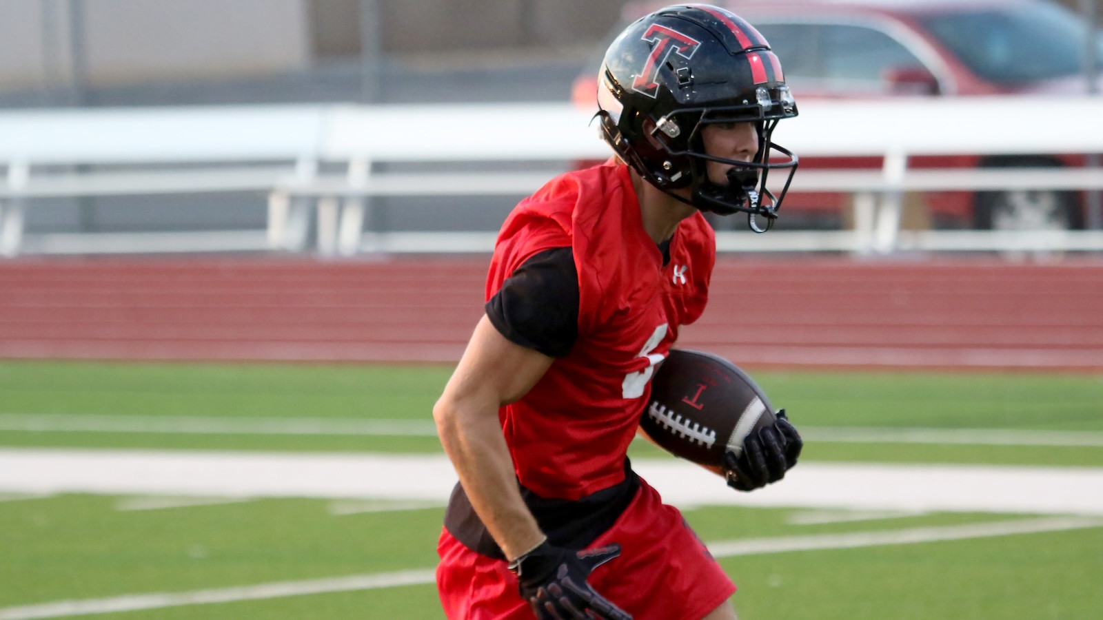 American football player in a red jersey and black helmet runs with a football on a green field.