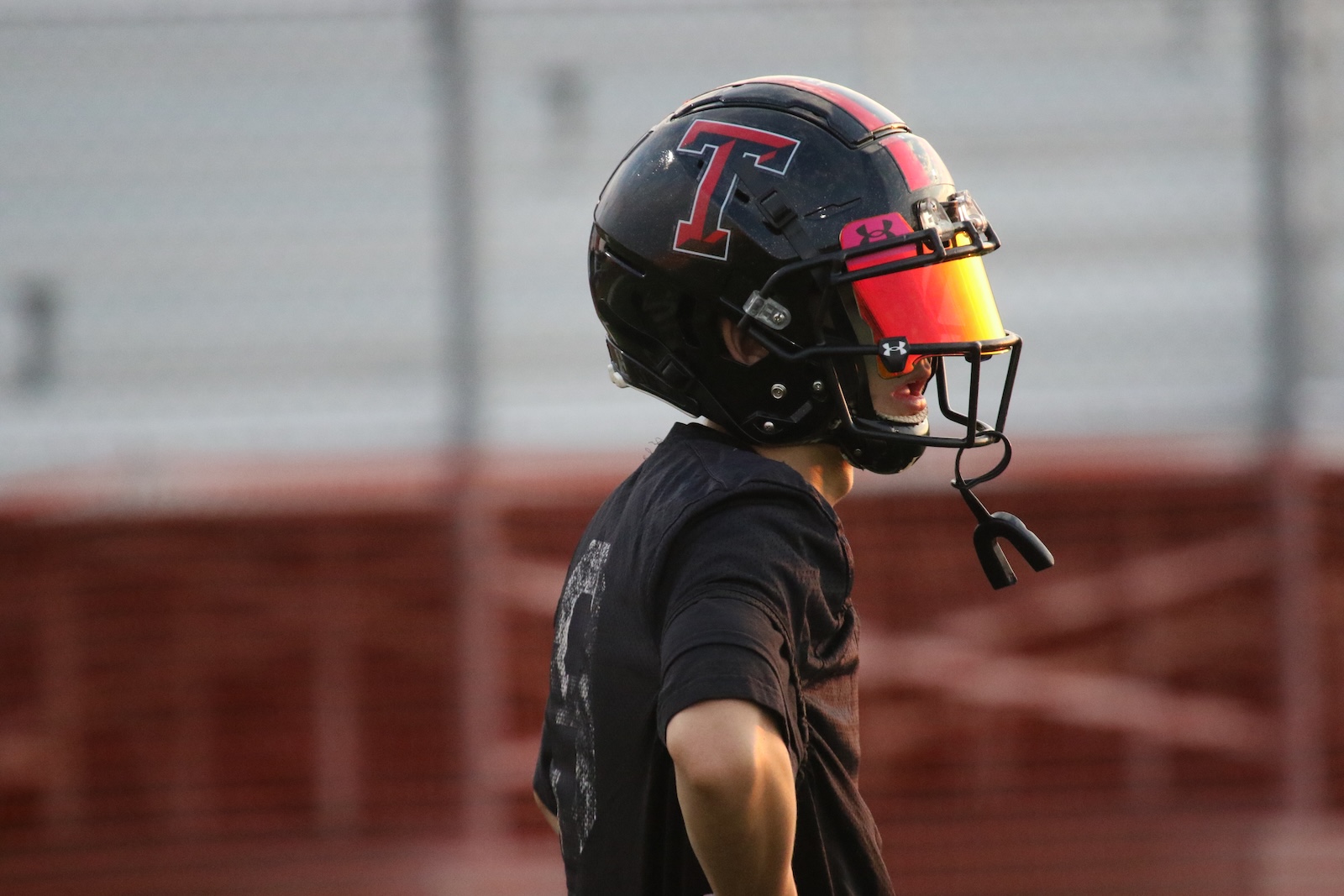 Side view of a football player wearing a black helmet with a red T logo and orange visor, standing on the field.