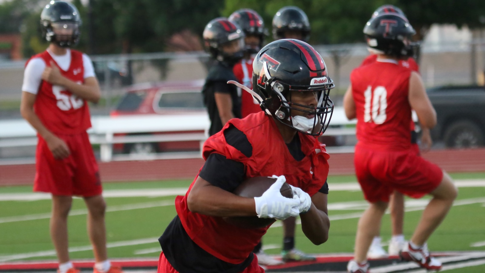 Football player in a red jersey runs with the ball during practice, wearing a black helmet and white gloves on a turf field.