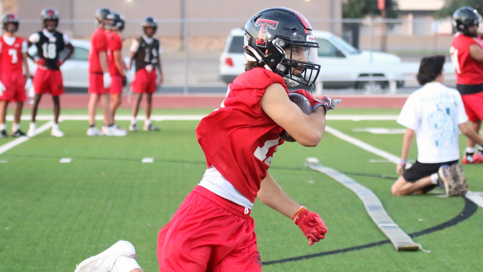 Youth football player in red jersey runs with the ball on artificial turf, teammates in red uniforms in the background.