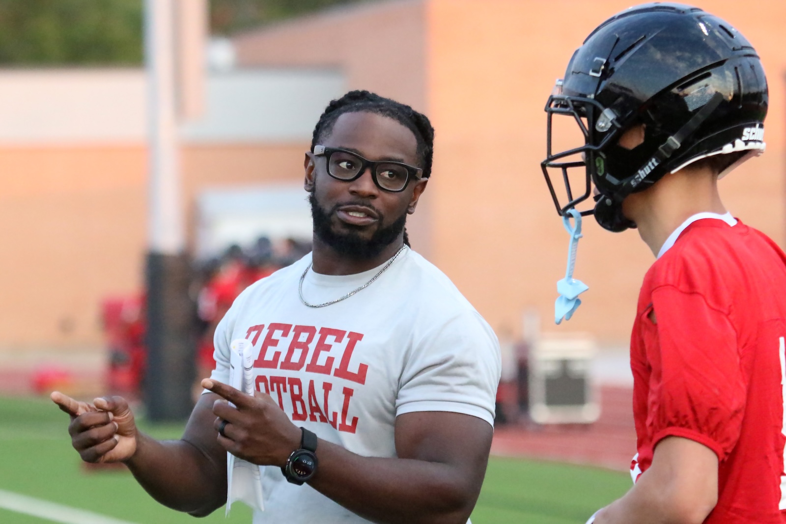 Coach with glasses and a beard talks to a football player in a red jersey on a practice field, pointing as he explains a drill.