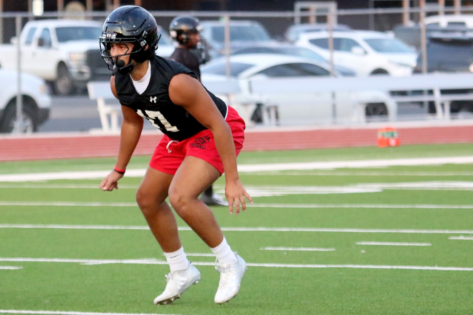 Youth football player in a black helmet and jersey, red shorts, crouched in a ready stance on a green artificial turf field.
