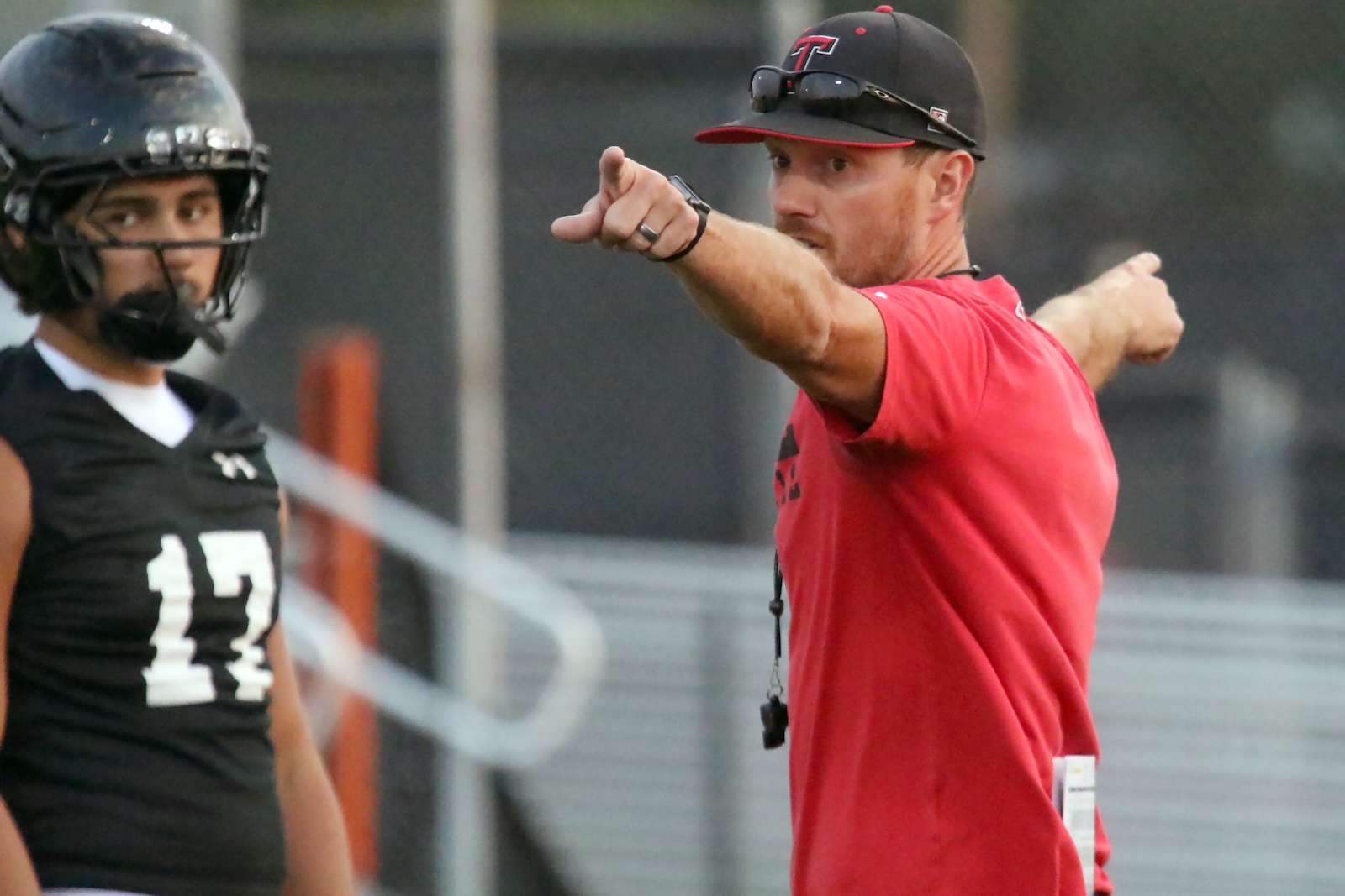 Coach in a red shirt and cap points emphatically while speaking to players on the sideline, with a football player in black uniform and helmet watching nearby.
