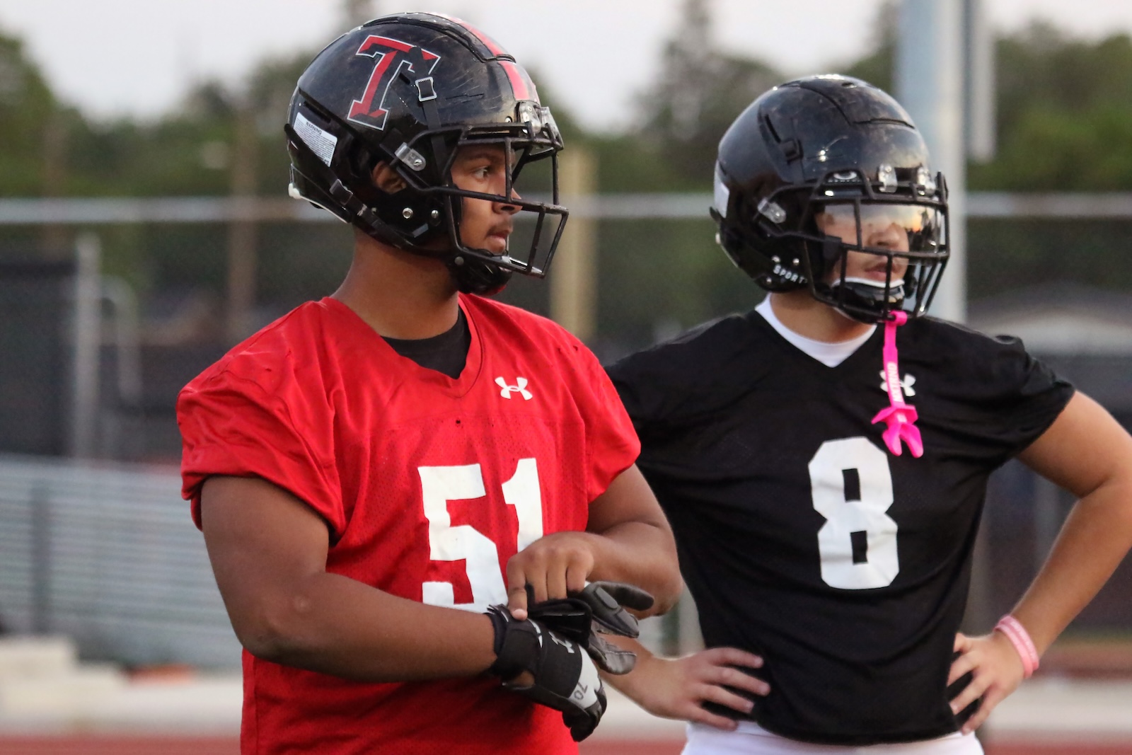 Two football players stand side by side on a field, one in a red jersey (#51) and the other in a black jersey (#8) wearing helmets and gloves, preparing for practice.