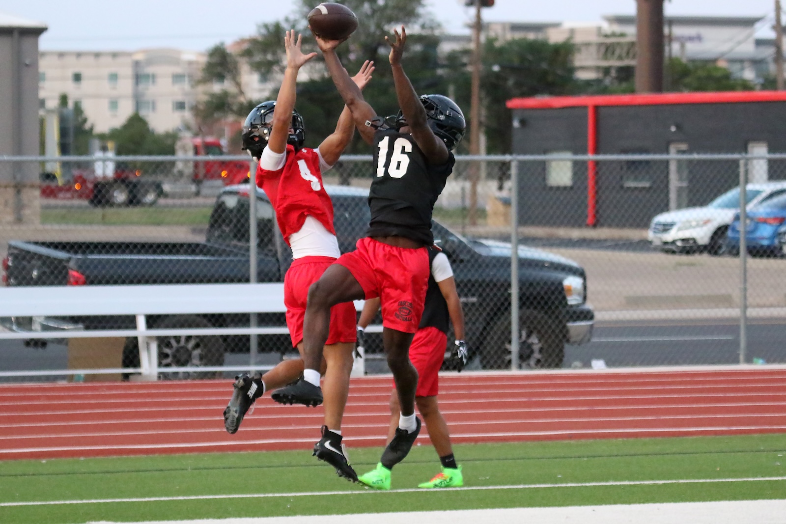 Two football players in red and black jump to catch a ball near the track, red #9 and black #16 forfeiting a high pass.