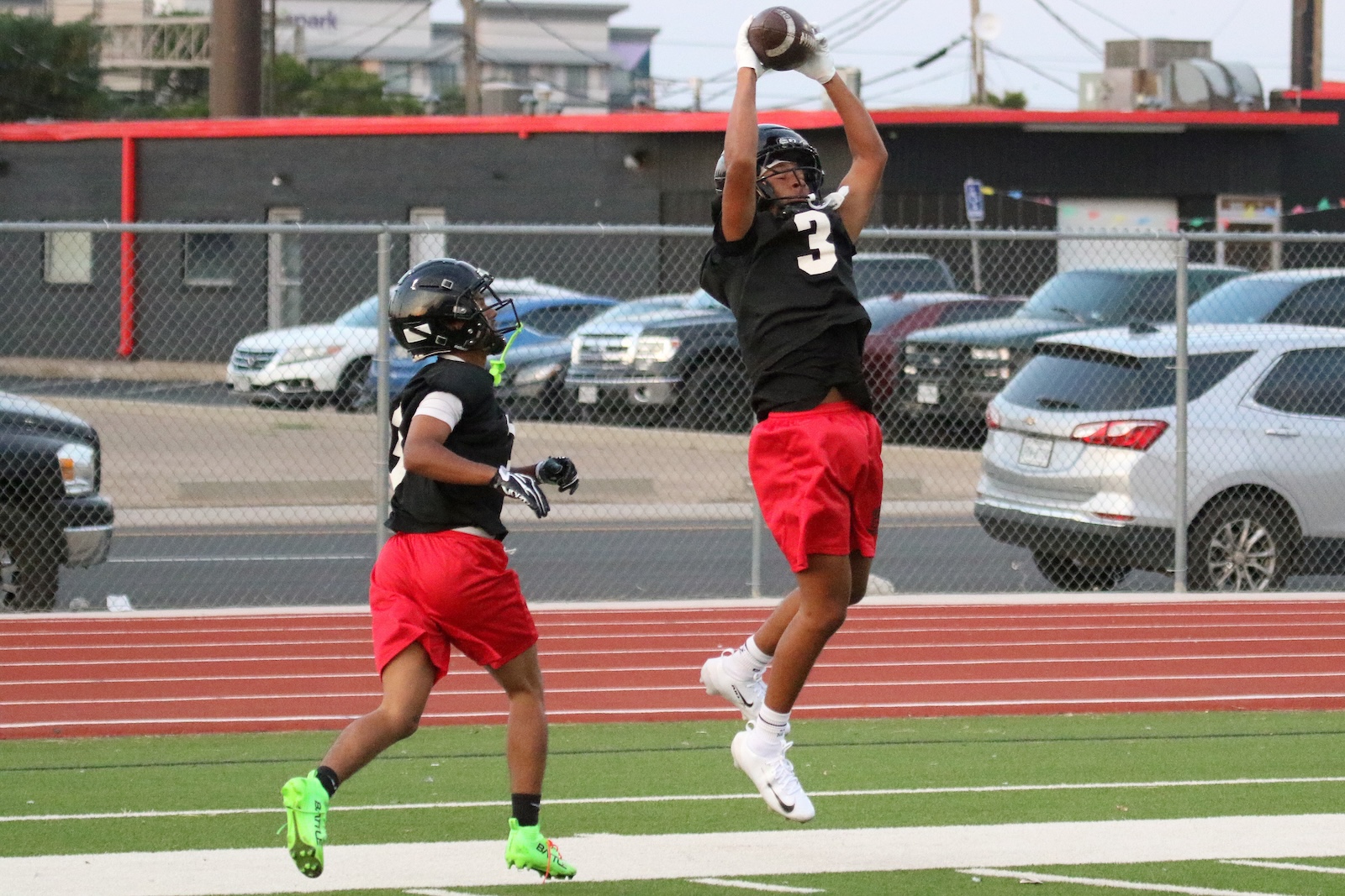 Two football players in black jerseys and red shorts compete for a ball on a track field; one leaps to catch a pass while the other pursues.
